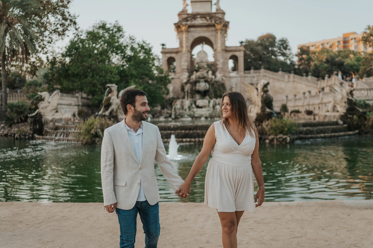 A hand-in-hand walk beside the fountain | Barcelona engagement photoshoot The couple walking hand in hand beside the green fountain pool, with the ornate monument behind them during their engagement photoshoot in Barcelona.