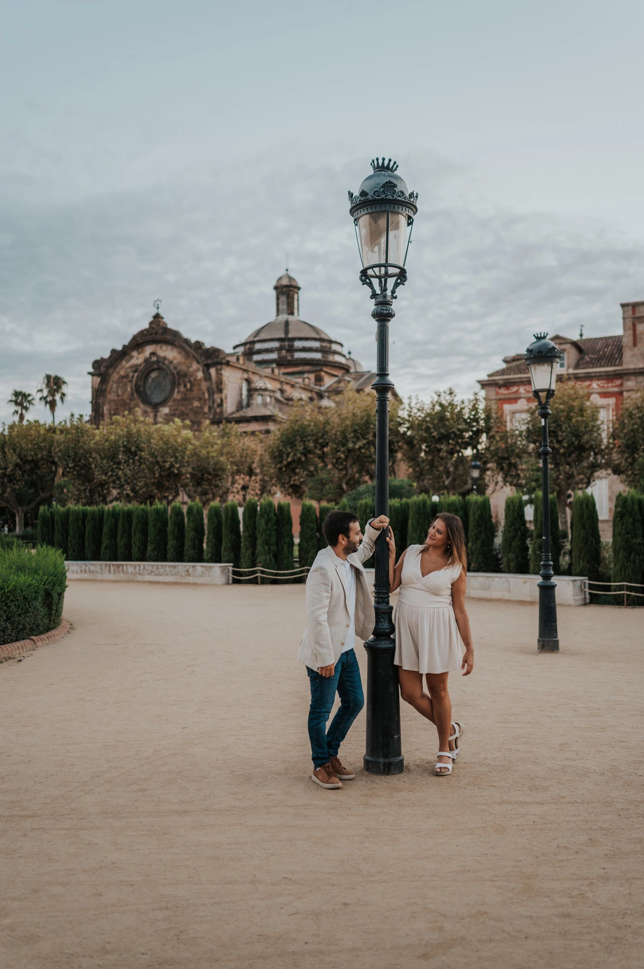 The couple standing together beside a tall lamppost, with clipped hedges and the park building behind them during their engagement photoshoot in Barcelona.
