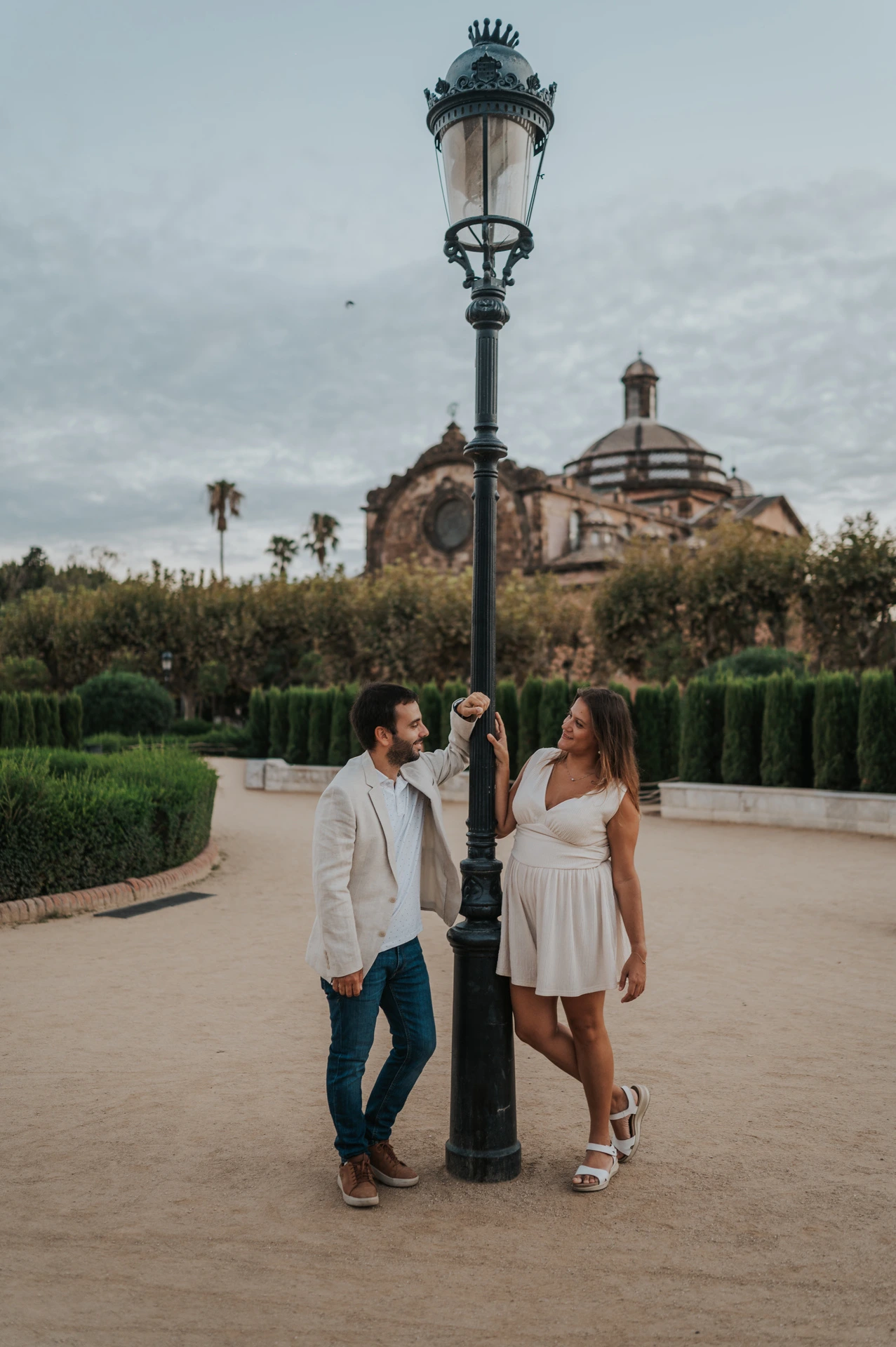 The couple standing together beside a tall lamppost, with clipped hedges and the park building behind them during their engagement photoshoot in Barcelona.