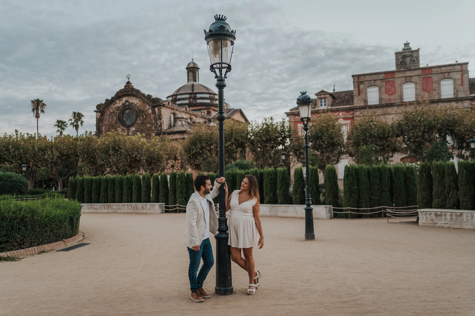 The couple standing together beside a tall lamppost, with clipped hedges and the park building behind them during their engagement photoshoot in Barcelona.