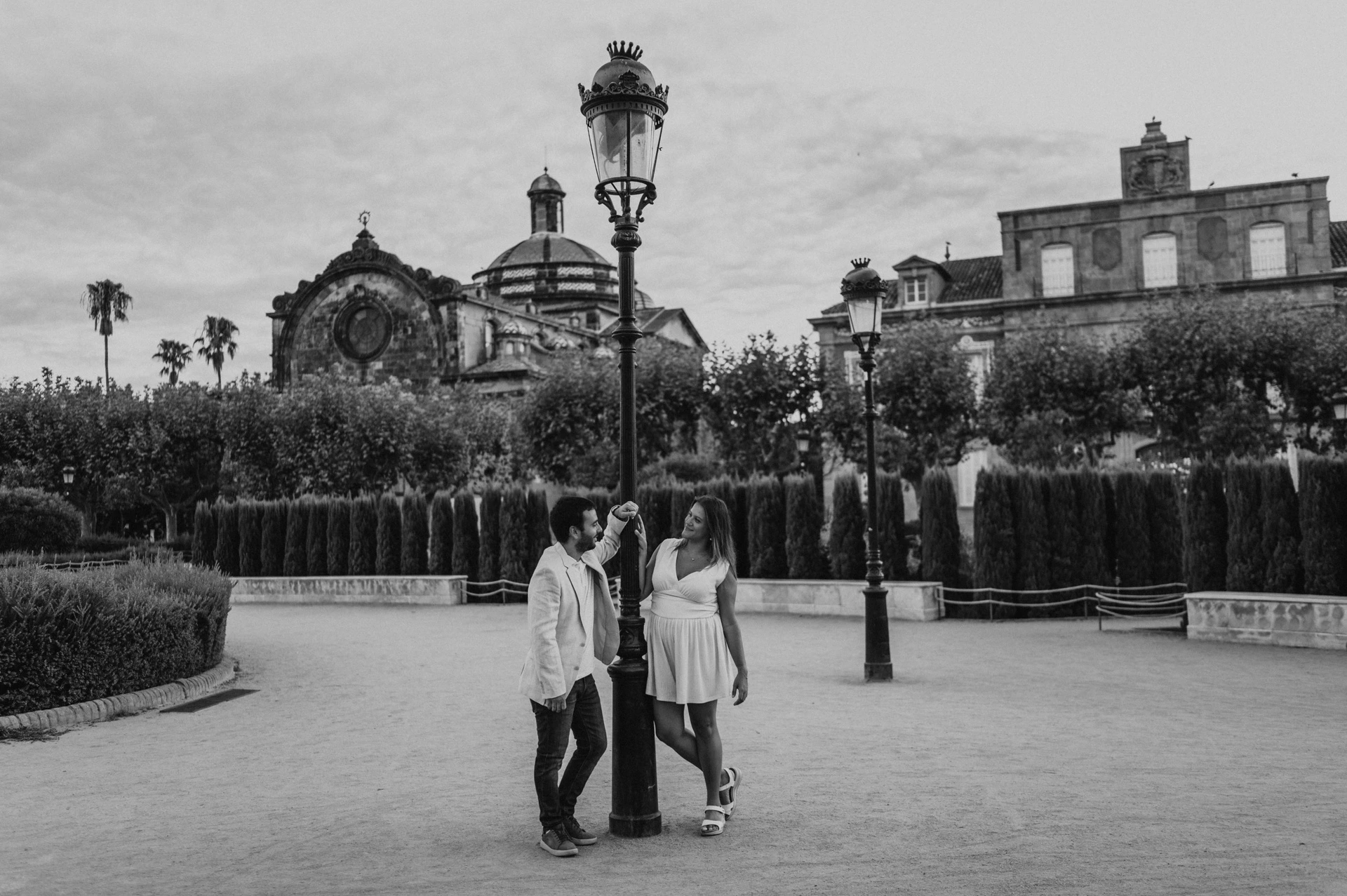 Black-and-white photograph: the couple standing together beside a tall lamppost, with clipped hedges and the park building behind them during their engagement photoshoot in Barcelona.