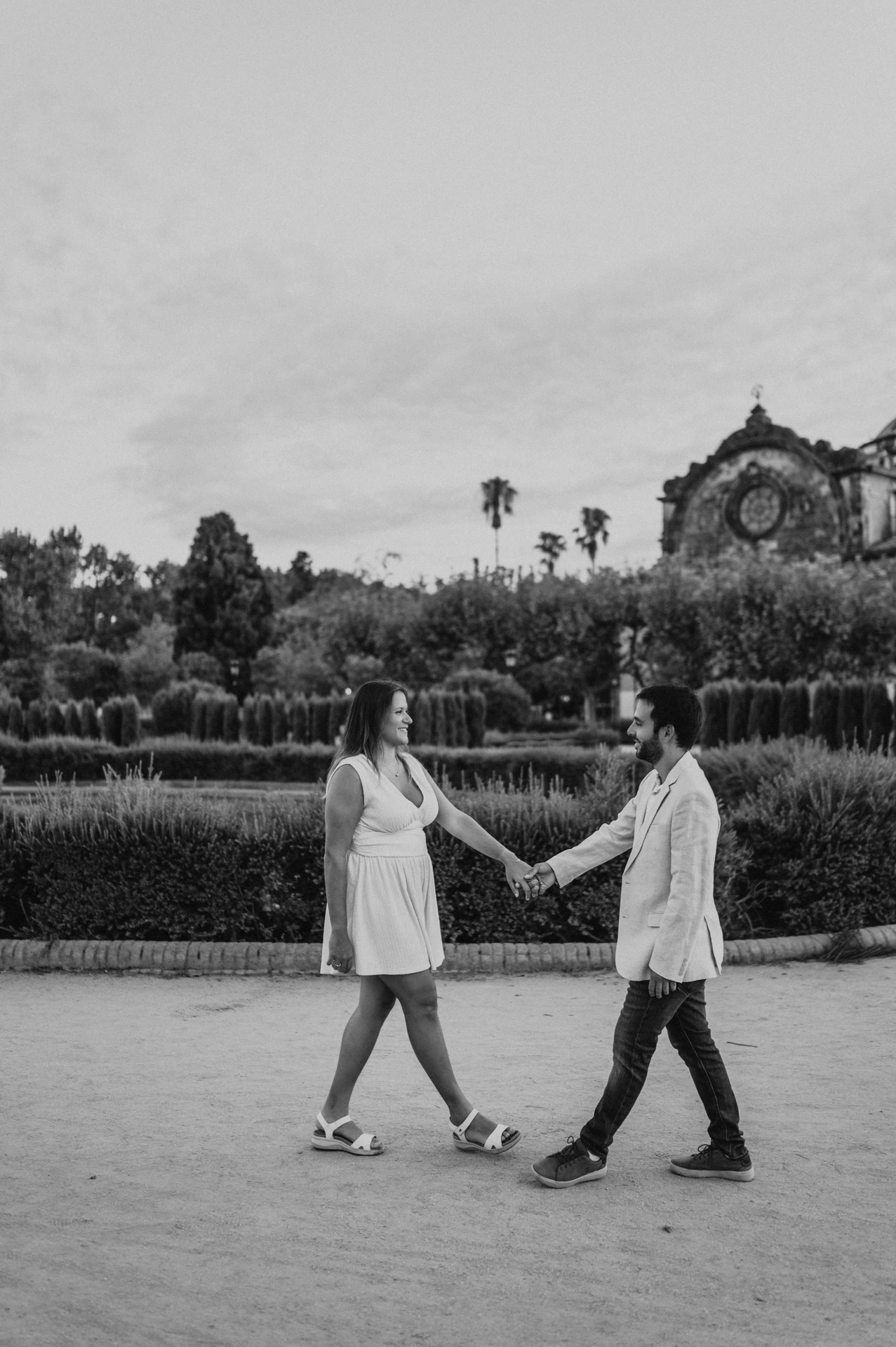 Black-and-white photograph: the couple facing each other and holding hands across a broad garden path during their engagement photoshoot in Barcelona.