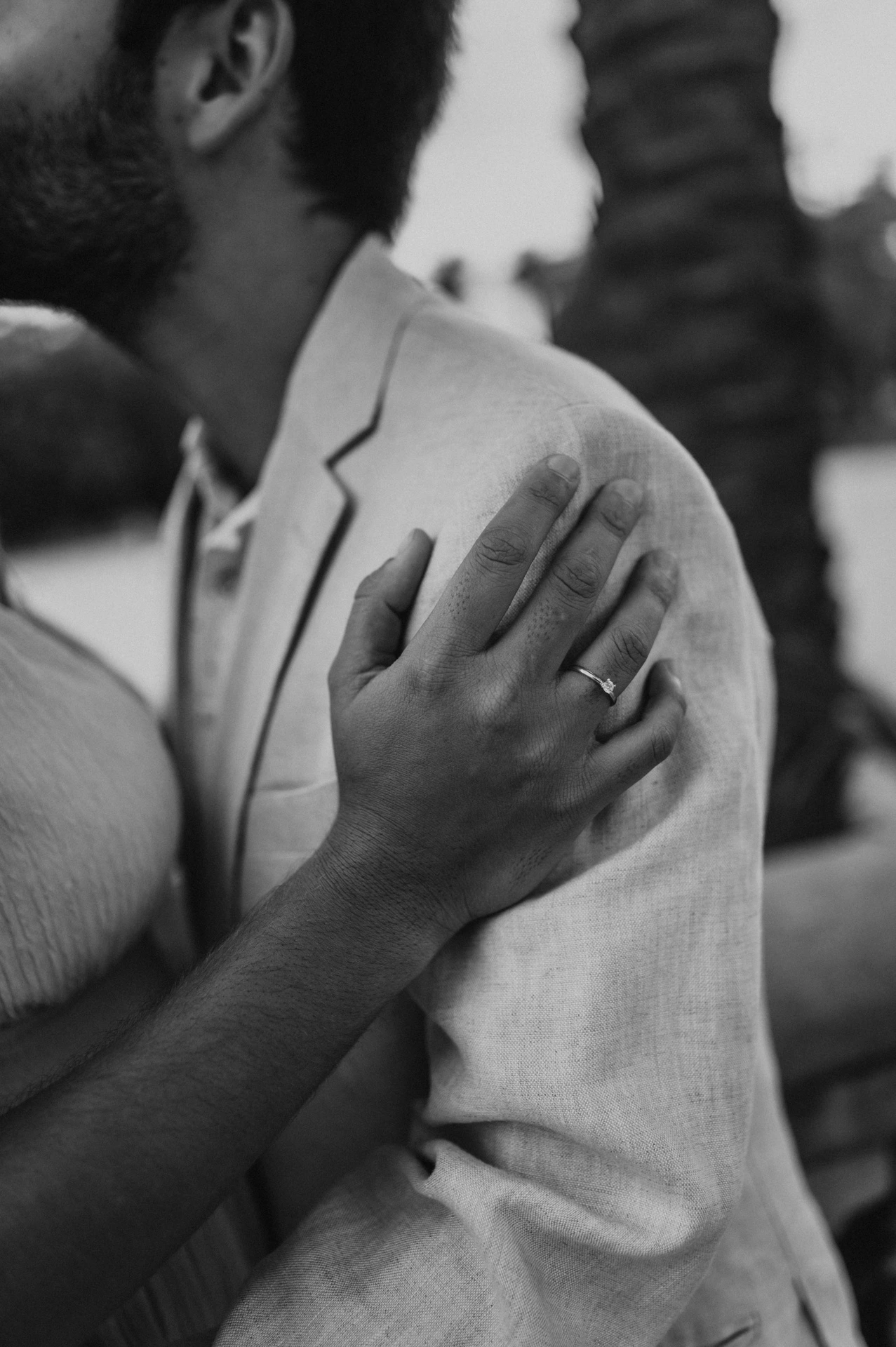 Black-and-white photograph: a close view of the woman’s hand resting on her partner’s light jacket as they hold each other during their engagement photoshoot in Barcelona.