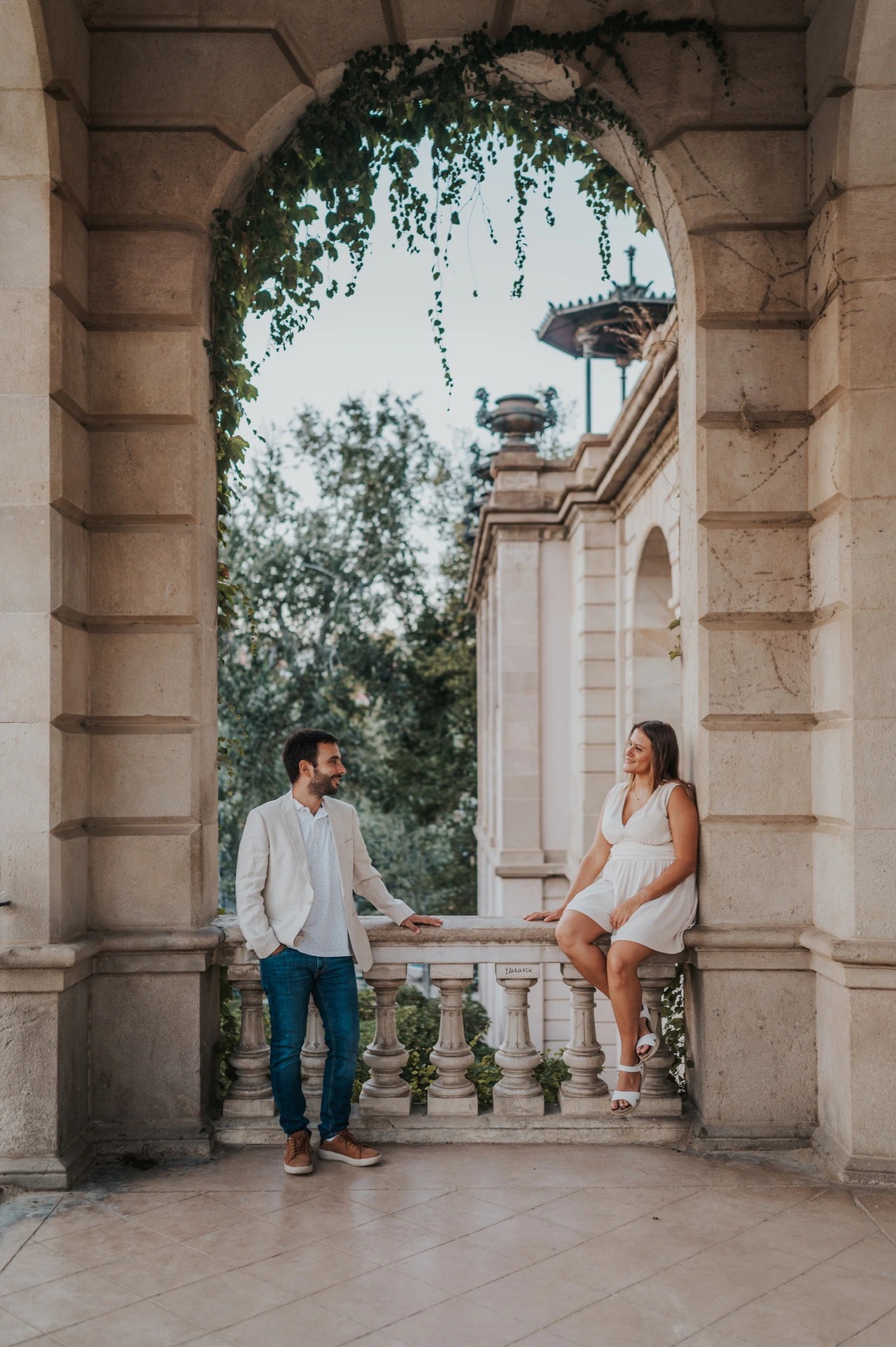 The woman sitting on a stone ledge beside her partner beneath an ivy-framed archway during their engagement photoshoot in Barcelona.