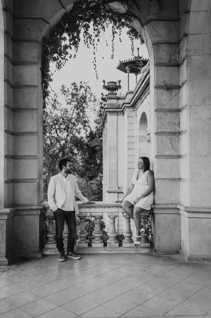 Together on a stone ledge beneath the ivy in black and white | Barcelona engagement photoshoot Black-and-white photograph: the woman sitting on a stone ledge beside her partner beneath an ivy-framed archway during their engagement photoshoot in Barcelona.