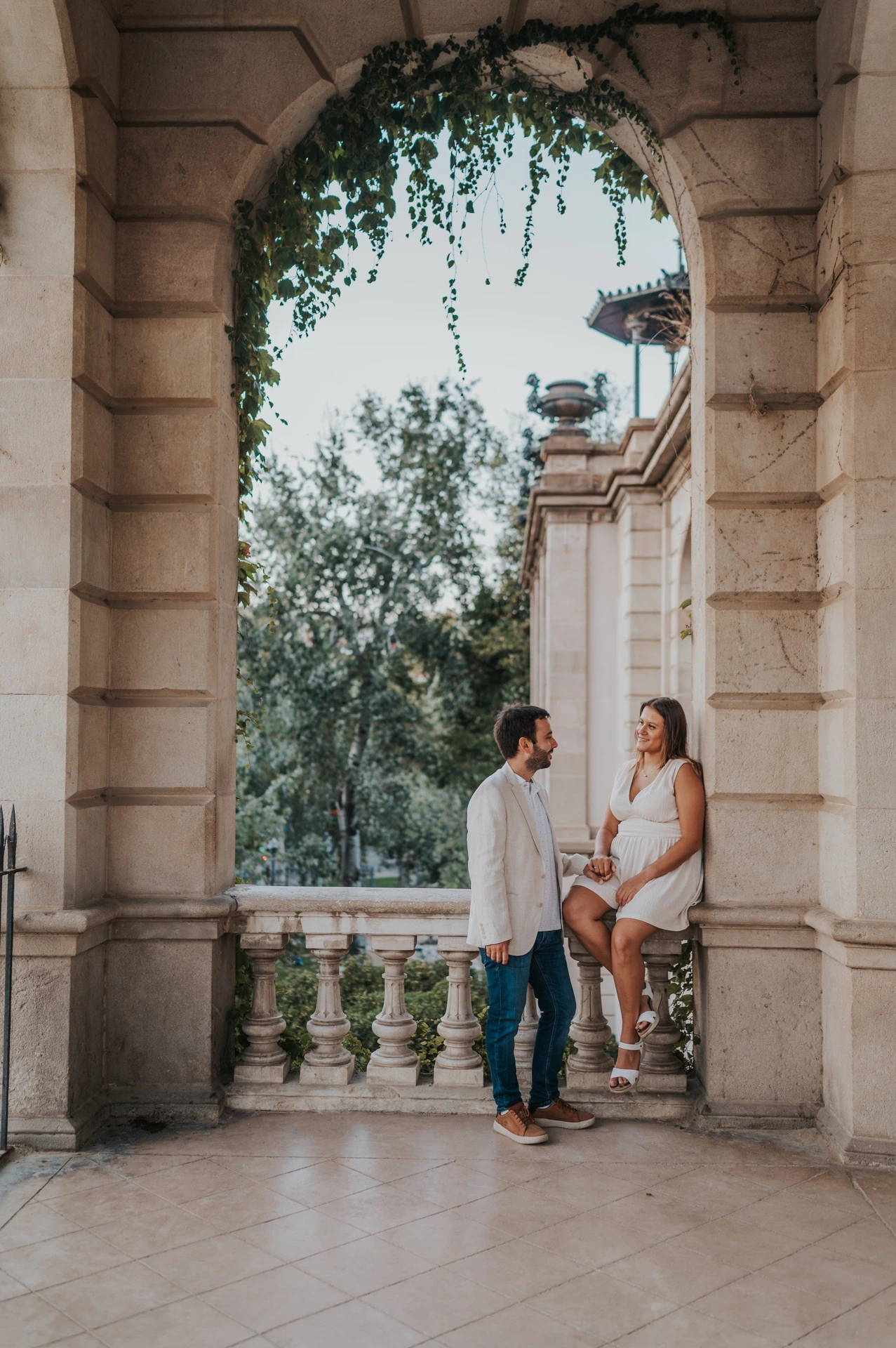 The woman sitting on a stone ledge beside her partner beneath an ivy-framed archway during their engagement photoshoot in Barcelona.