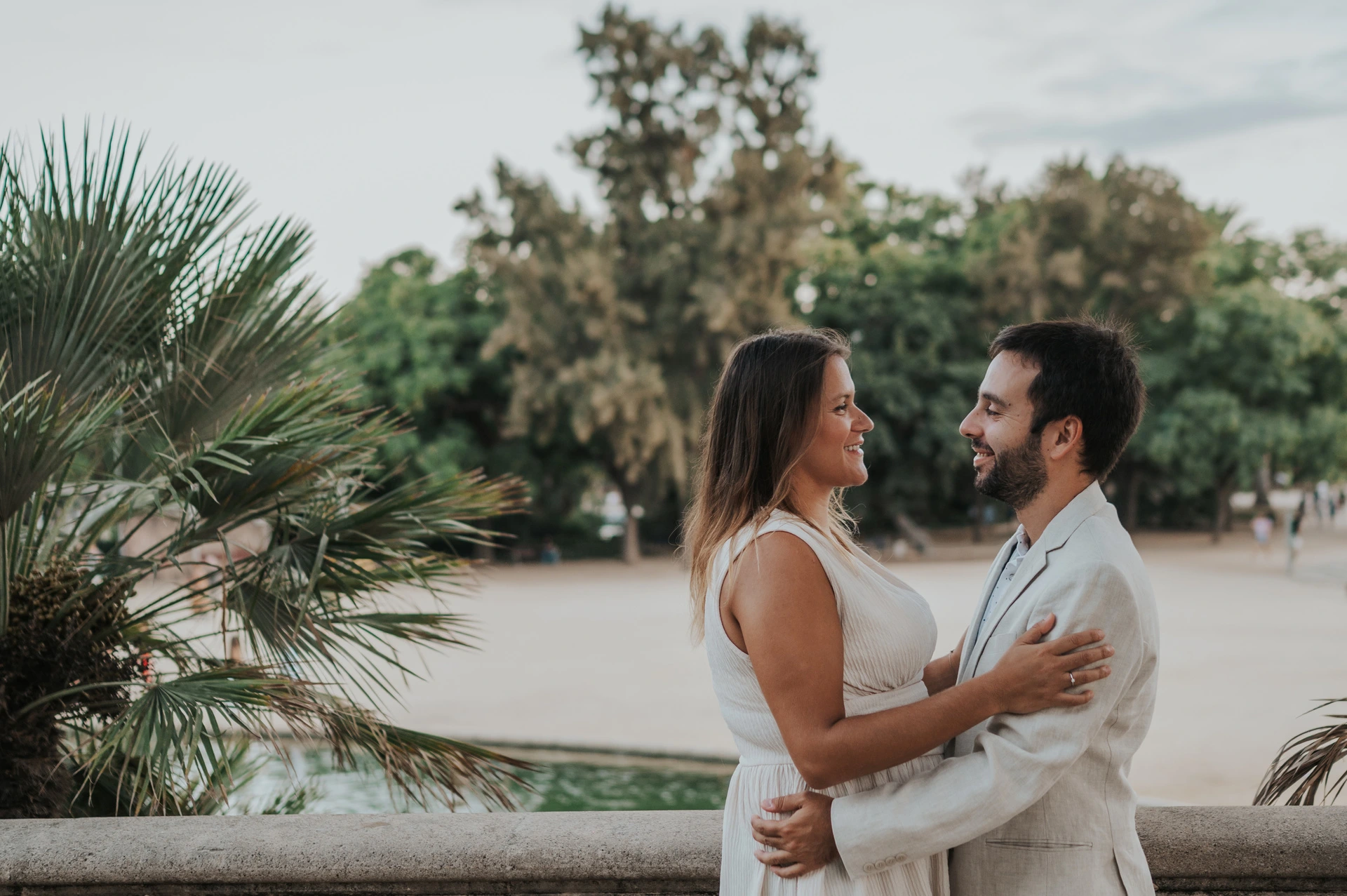 The couple holding each other beside the terrace balustrade with palms in the background during their engagement photoshoot in Barcelona.