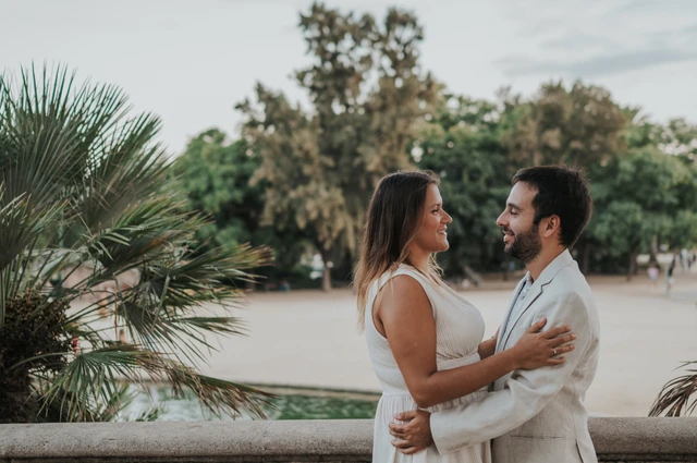 The couple holding each other beside the terrace balustrade with palms in the background during their engagement photoshoot in Barcelona.