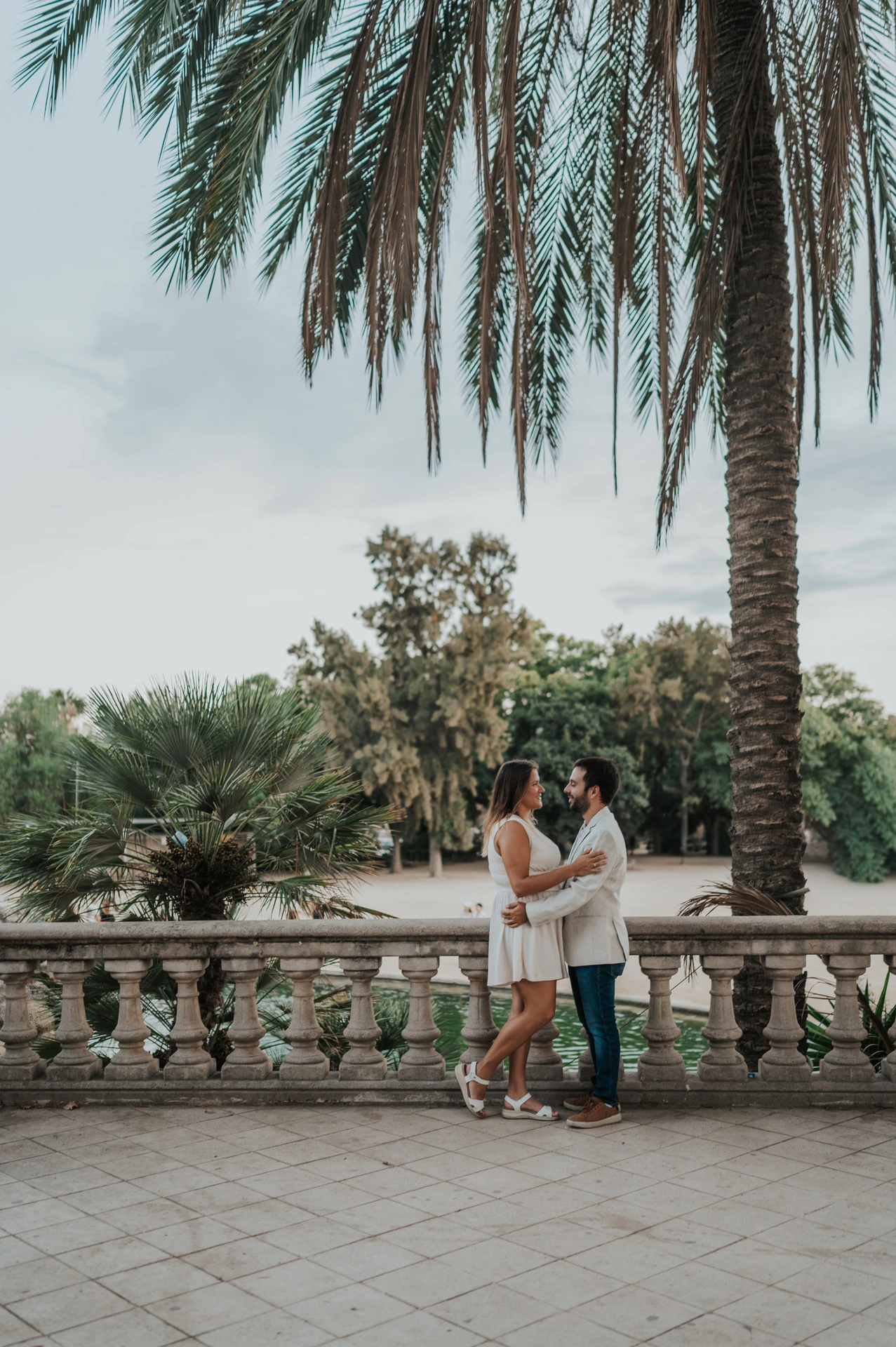 The couple holding each other beside the terrace balustrade with palms in the background during their engagement photoshoot in Barcelona.