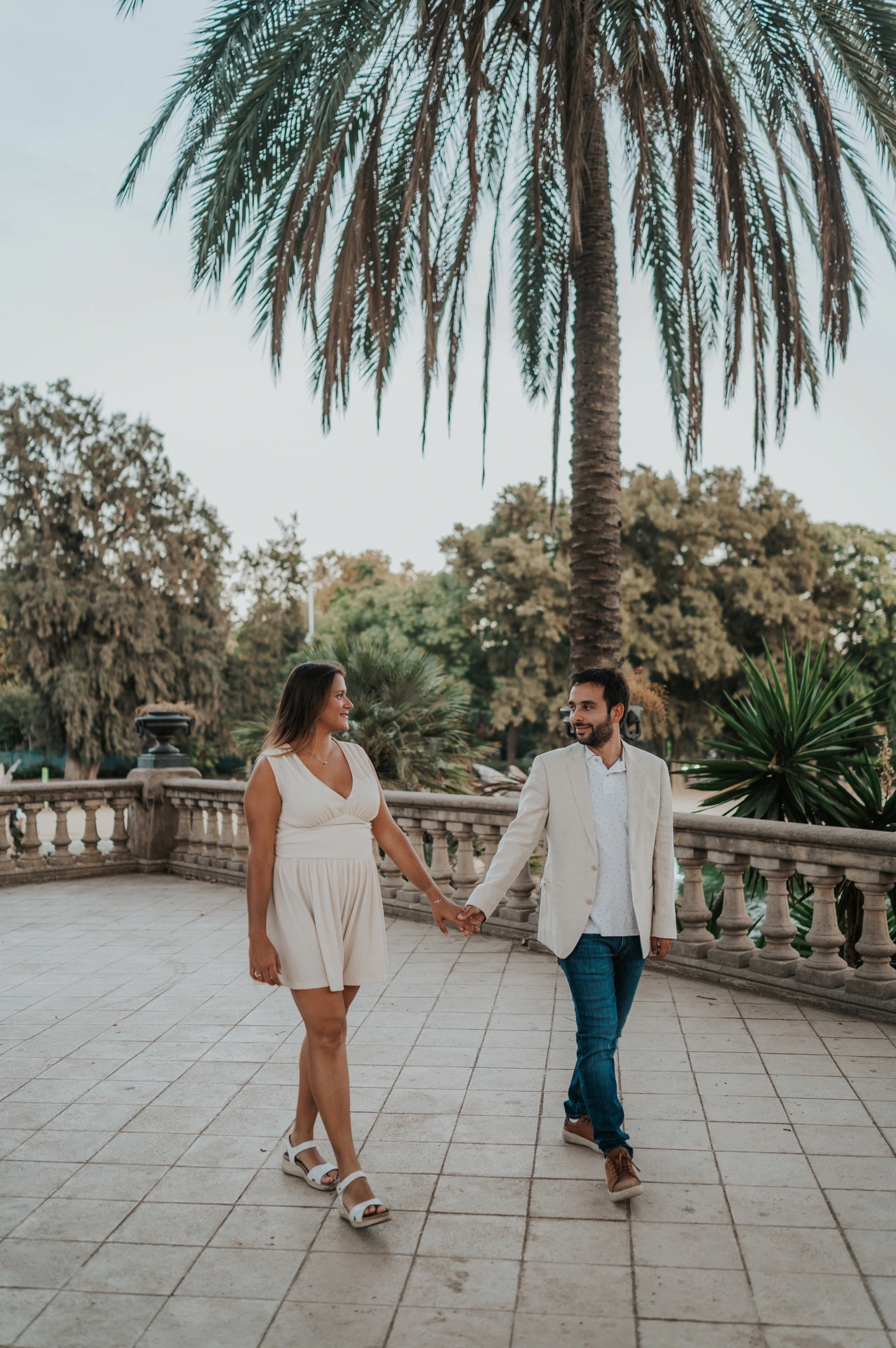 The couple walking hand in hand across a terrace beside a palm tree and stone railing during their engagement photoshoot in Barcelona.