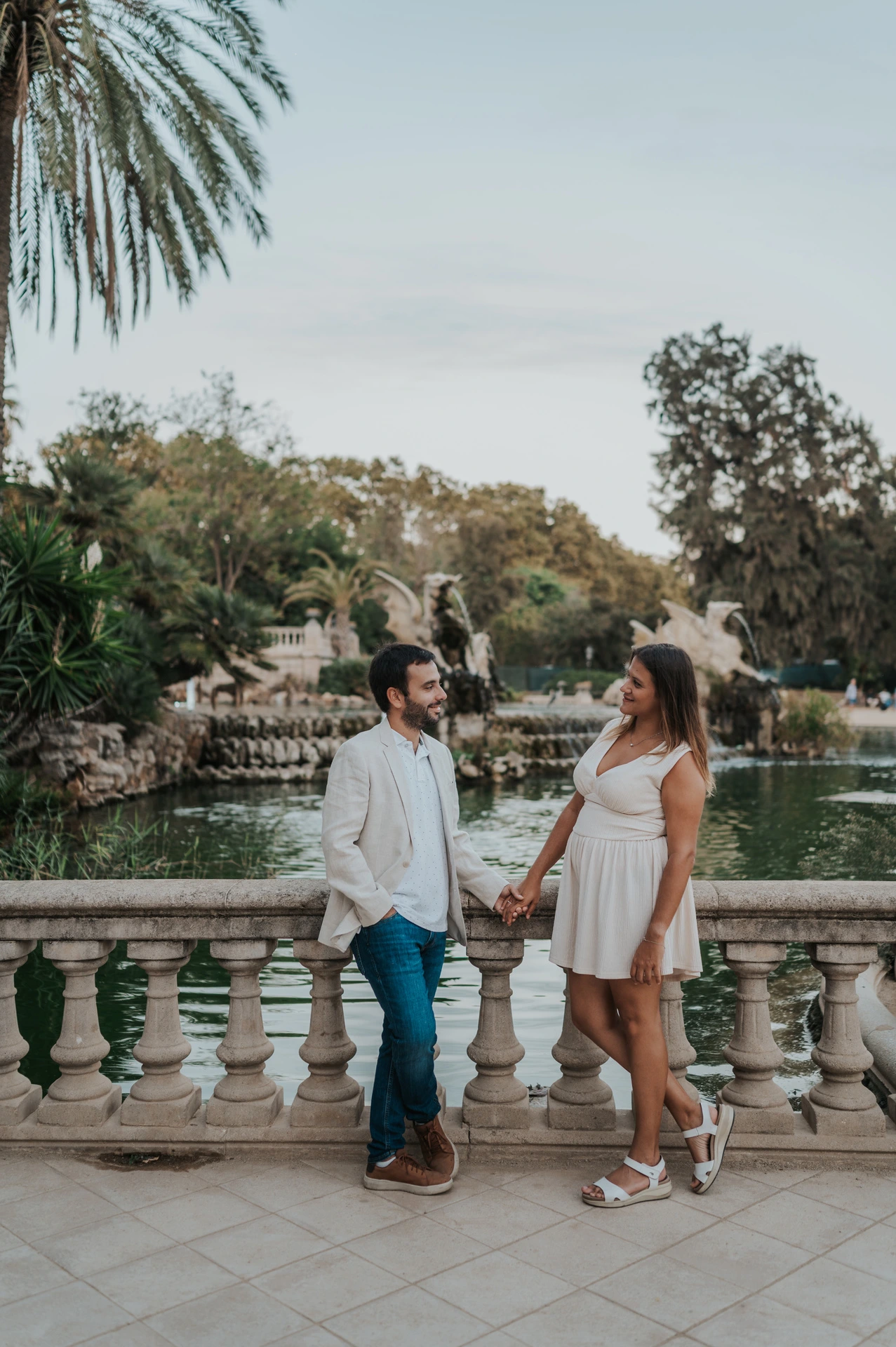The couple walking hand in hand across a terrace beside a palm tree and stone railing during their engagement photoshoot in Barcelona.