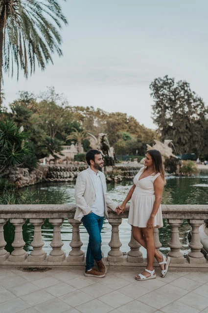 A walk along the palm-lined terrace | Barcelona engagement photoshoot The couple walking hand in hand across a terrace beside a palm tree and stone railing during their engagement photoshoot in Barcelona.