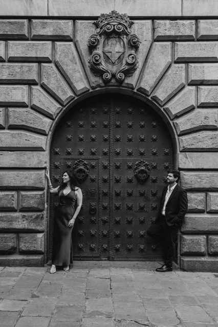 Portraits beside an arched wooden door in black and white | Barcelona engagement photoshoot Black-and-white photograph: the couple standing on opposite sides of a large studded wooden door in an arched stone entrance in the Gothic Quarter during their engagement photoshoot in Barcelona.