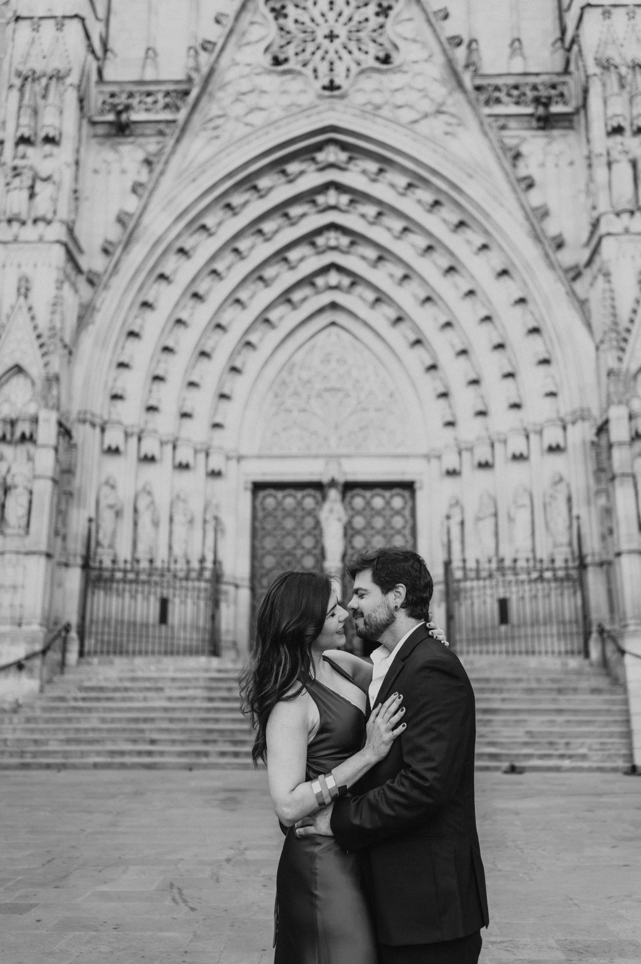 Black-and-white photograph: the couple standing close together in front of Barcelona Cathedral’s carved Gothic entrance during their engagement photoshoot in Barcelona.