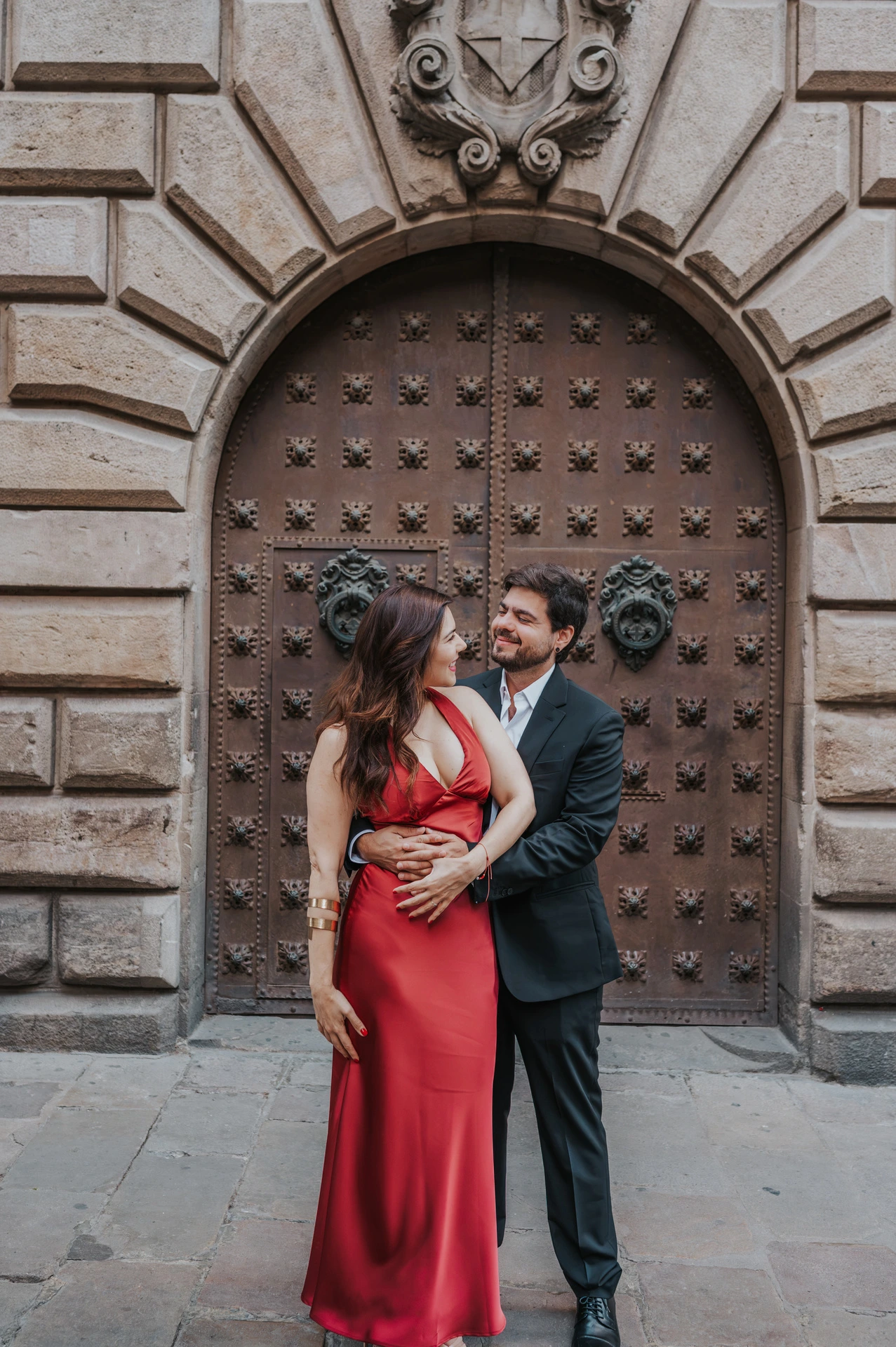 The man embracing his partner from behind in front of a studded wooden door in the Gothic Quarter during their engagement photoshoot in Barcelona.