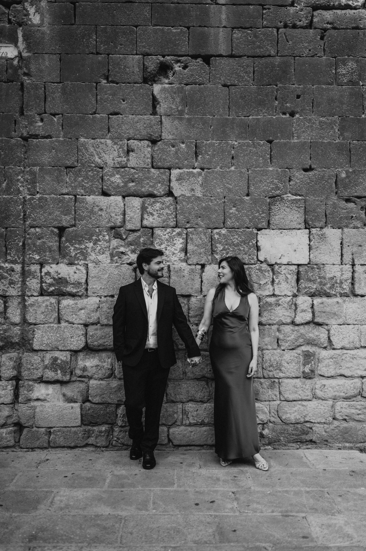 Black-and-white photograph: the couple standing together against a textured historic stone wall in the Gothic Quarter during their engagement photoshoot in Barcelona.