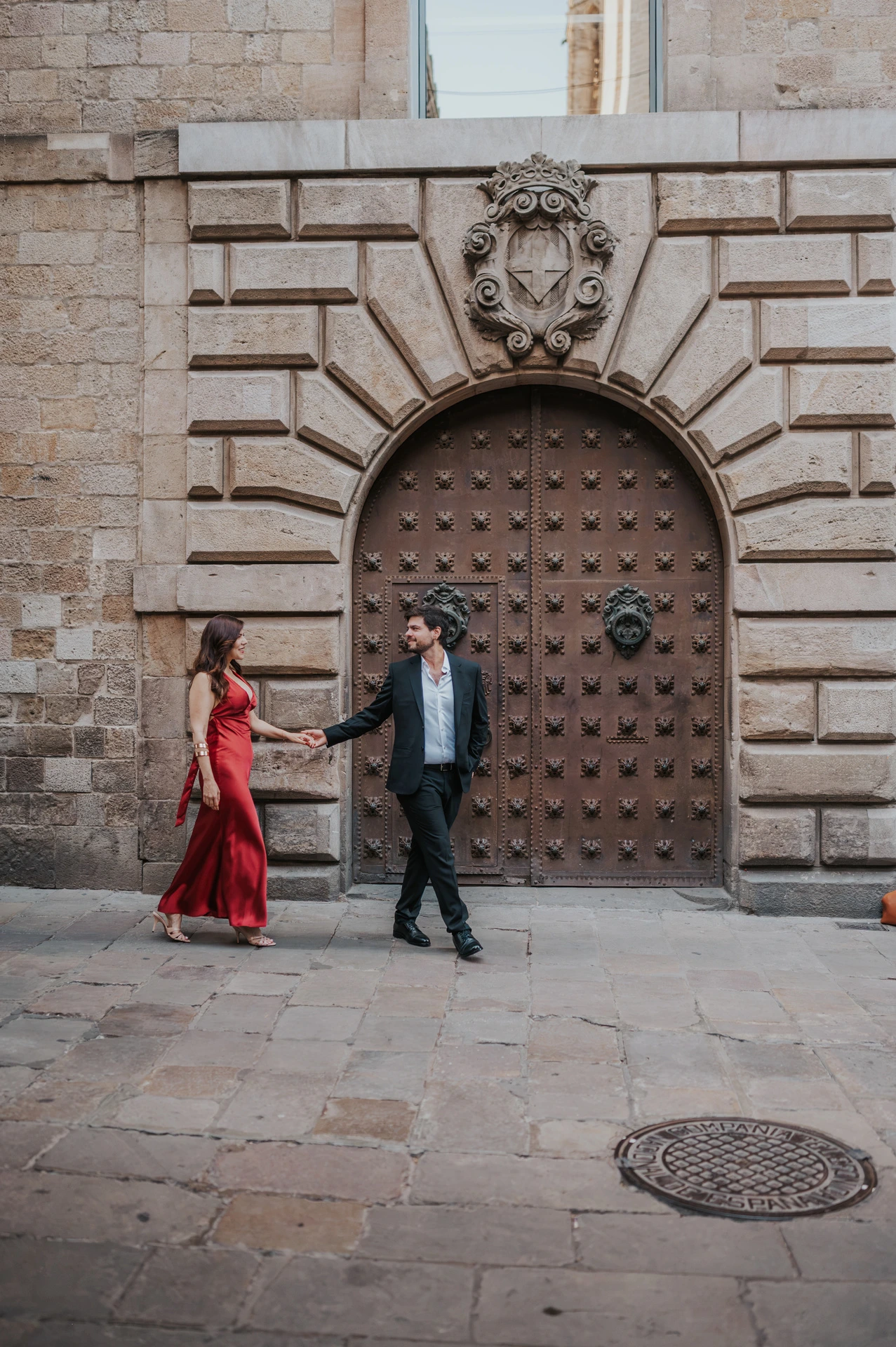 The couple holding hands in front of a large arched wooden door in the Gothic Quarter during their engagement photoshoot in Barcelona.