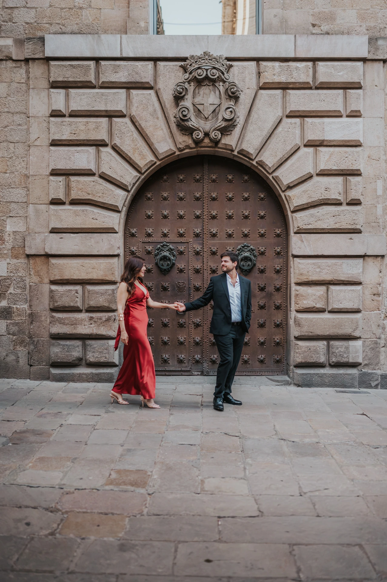 The couple holding hands in front of a large arched wooden door in the Gothic Quarter during their engagement photoshoot in Barcelona.
