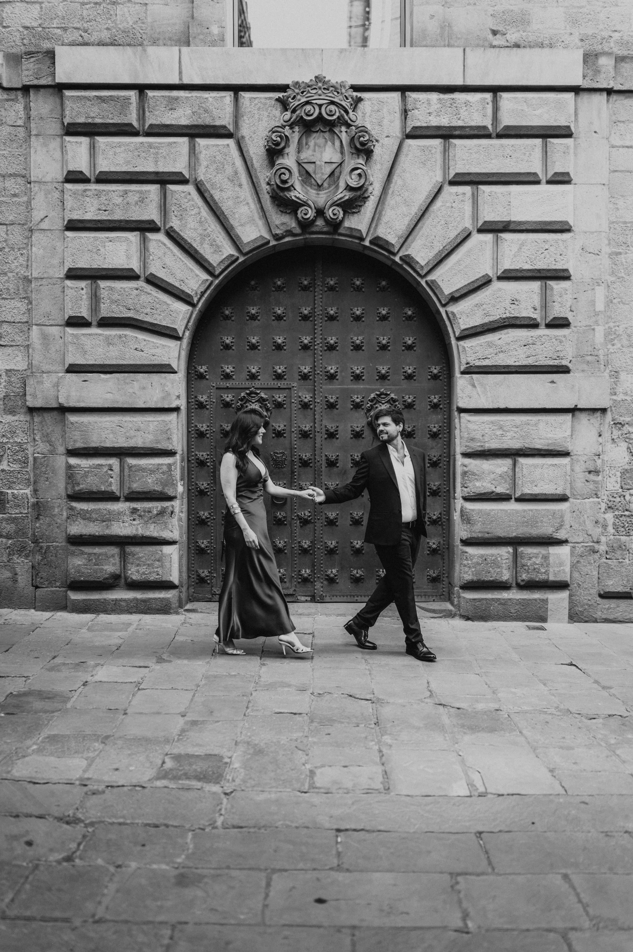Black-and-white photograph: the couple holding hands in front of a large arched wooden door in the Gothic Quarter during their engagement photoshoot in Barcelona.