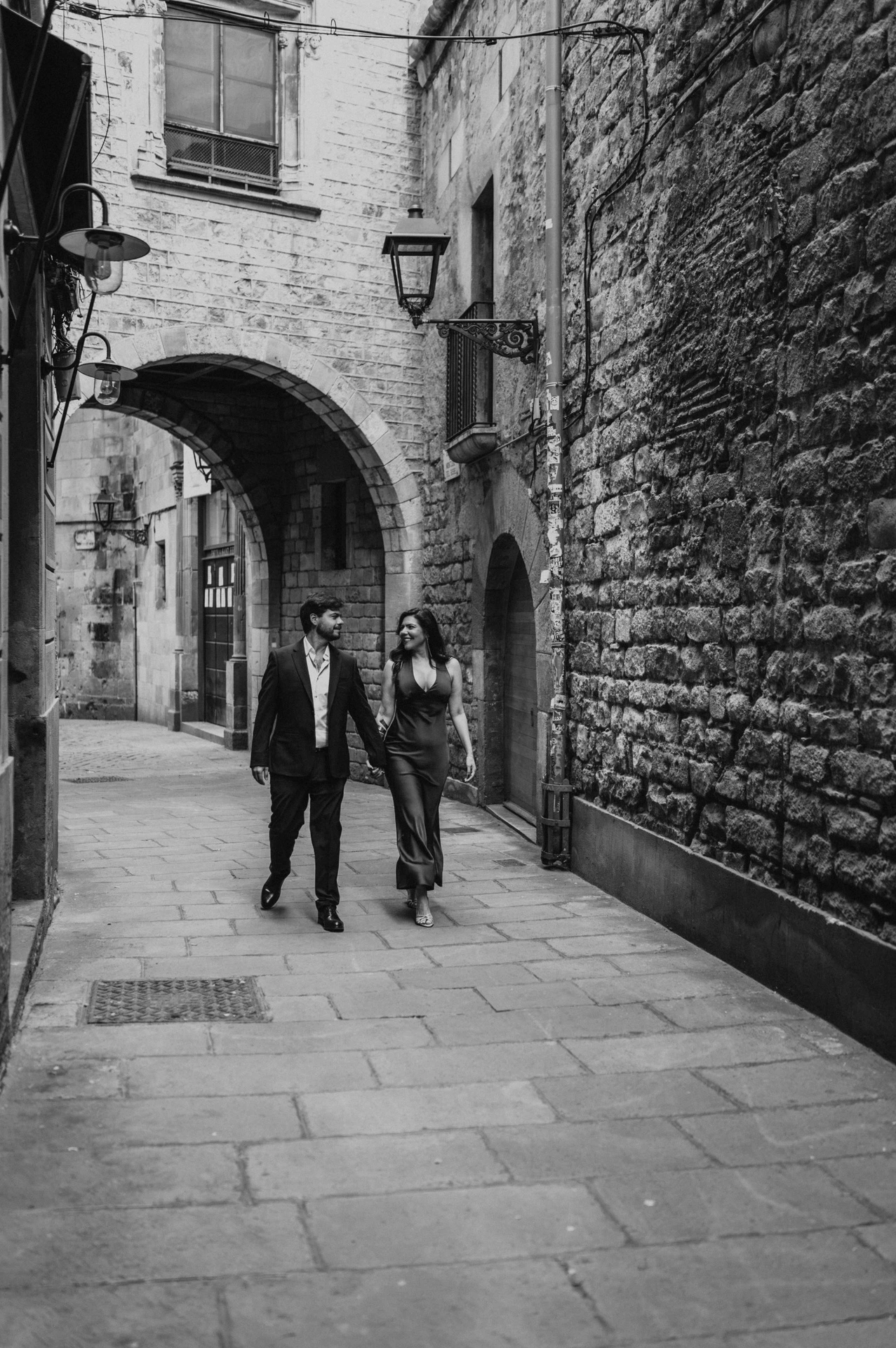 Black-and-white photograph: the couple walking together through a stone lane beneath an arch in the Gothic Quarter during their engagement photoshoot in Barcelona.
