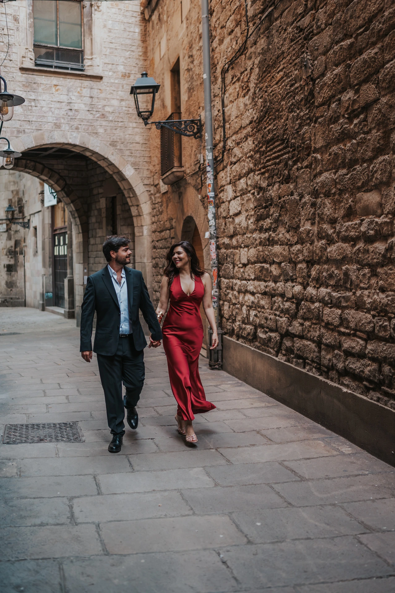 The couple walking together through a stone lane beneath an arch in the Gothic Quarter during their engagement photoshoot in Barcelona.