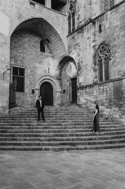 The architecture of Plaça del Rei around them in black and white | Barcelona engagement photoshoot Black-and-white photograph: the couple standing on the broad stone steps at Plaça del Rei, surrounded by tall stone walls, arches and historic windows during their engagement photoshoot in Barcelona.