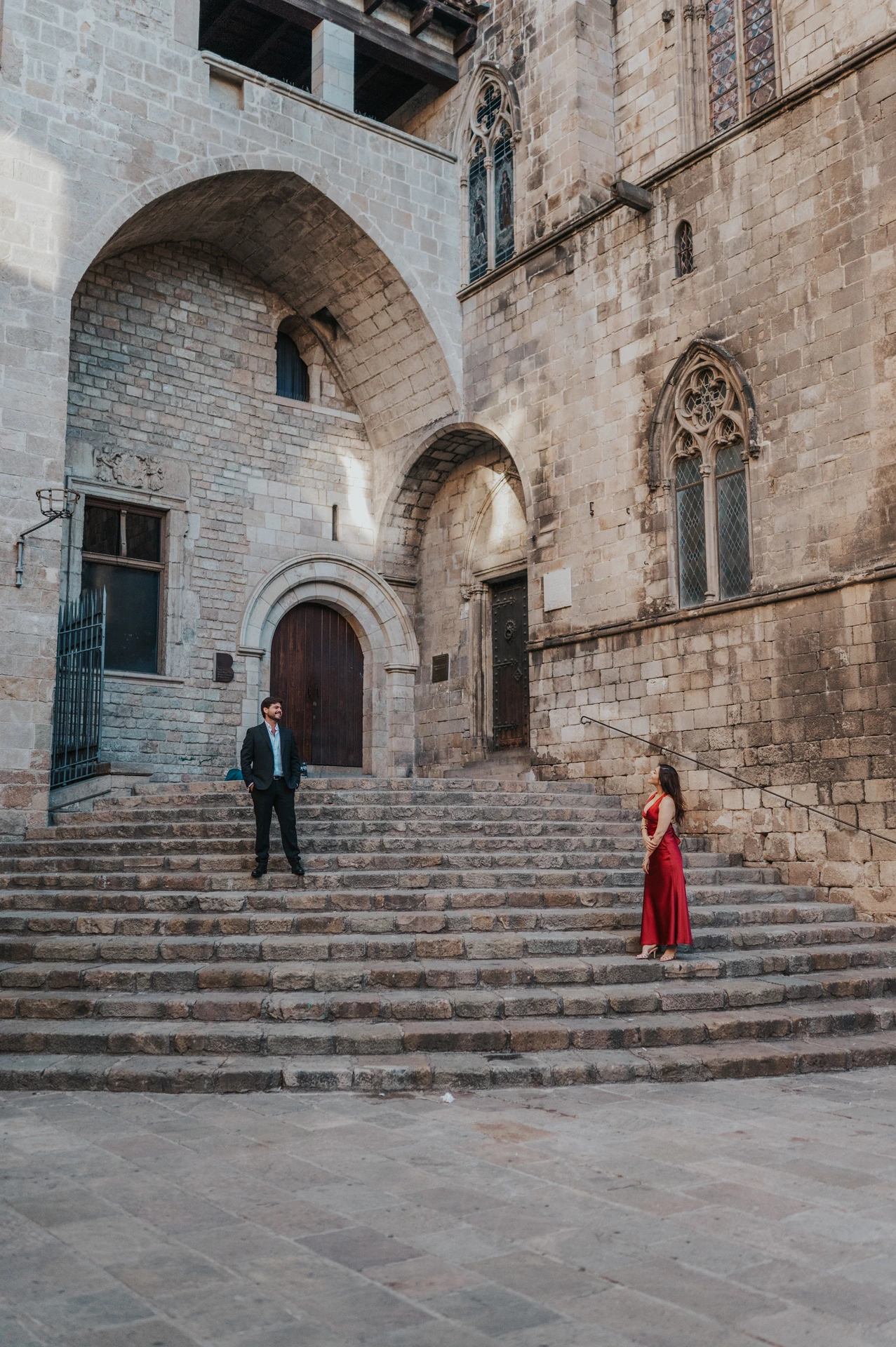 The couple standing on the broad stone steps at Plaça del Rei, surrounded by tall stone walls, arches and historic windows during their engagement photoshoot in Barcelona.