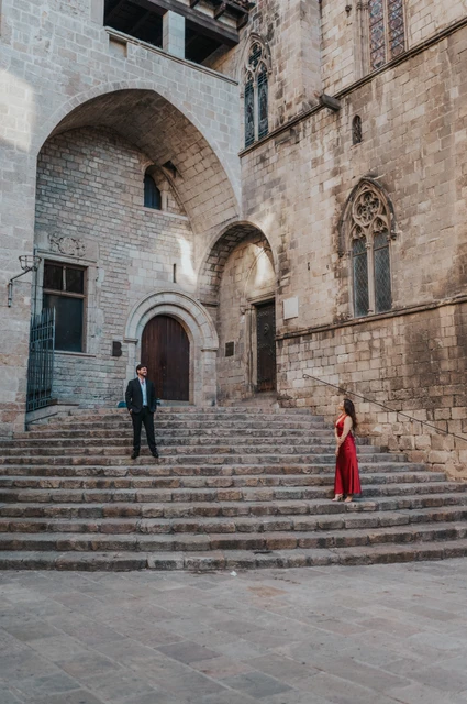 The architecture of Plaça del Rei around them | Barcelona engagement photoshoot The couple standing on the broad stone steps at Plaça del Rei, surrounded by tall stone walls, arches and historic windows during their engagement photoshoot in Barcelona.