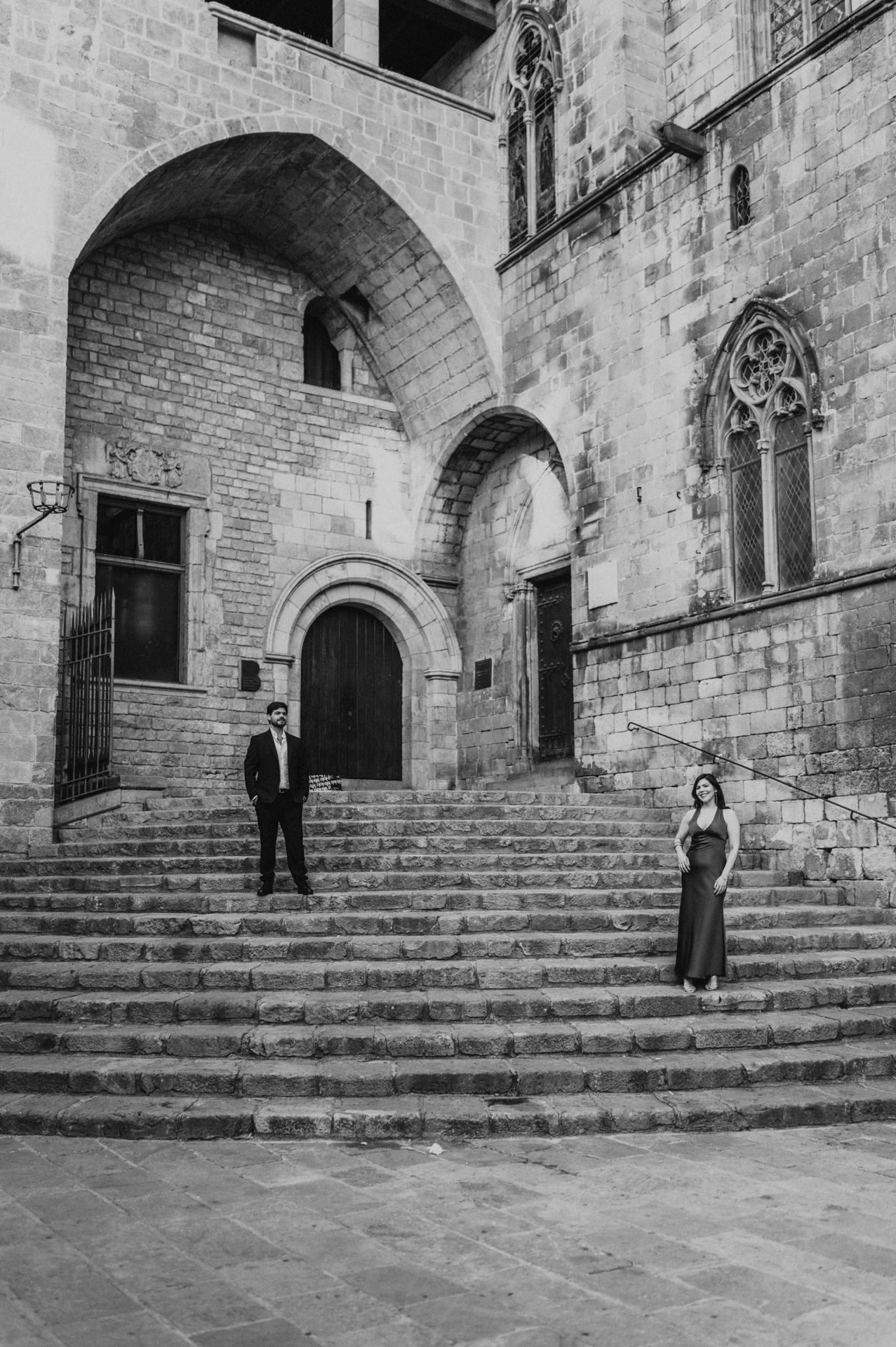 Black-and-white photograph: the couple standing on the broad stone steps at Plaça del Rei, surrounded by tall stone walls, arches and historic windows during their engagement photoshoot in Barcelona.
