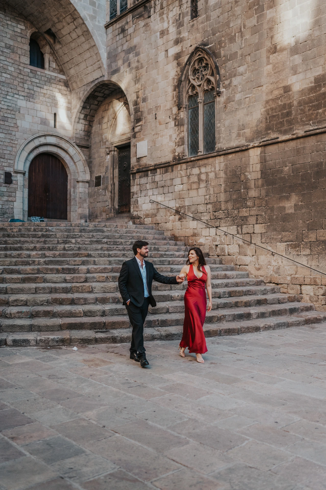 The couple walking together across the square below the broad stone steps of Plaça del Rei during their engagement photoshoot in Barcelona.