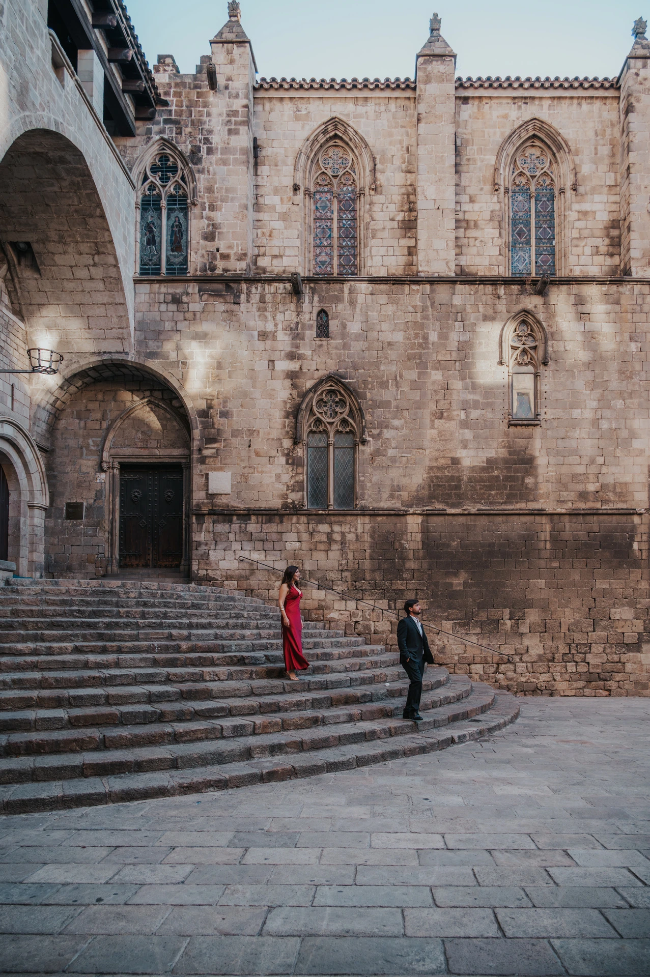 The couple holding hands on the broad stone steps at Plaça del Rei, with historic doors and pointed windows behind them during their engagement photoshoot in Barcelona.