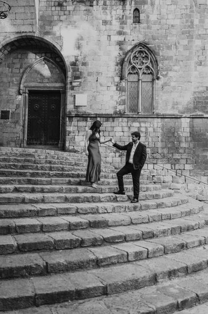 Together on the stone steps of Plaça del Rei in black and white | Barcelona engagement photoshoot Black-and-white photograph: the couple holding hands on the broad stone steps at Plaça del Rei, with historic doors and pointed windows behind them during their engagement photoshoot in Barcelona.
