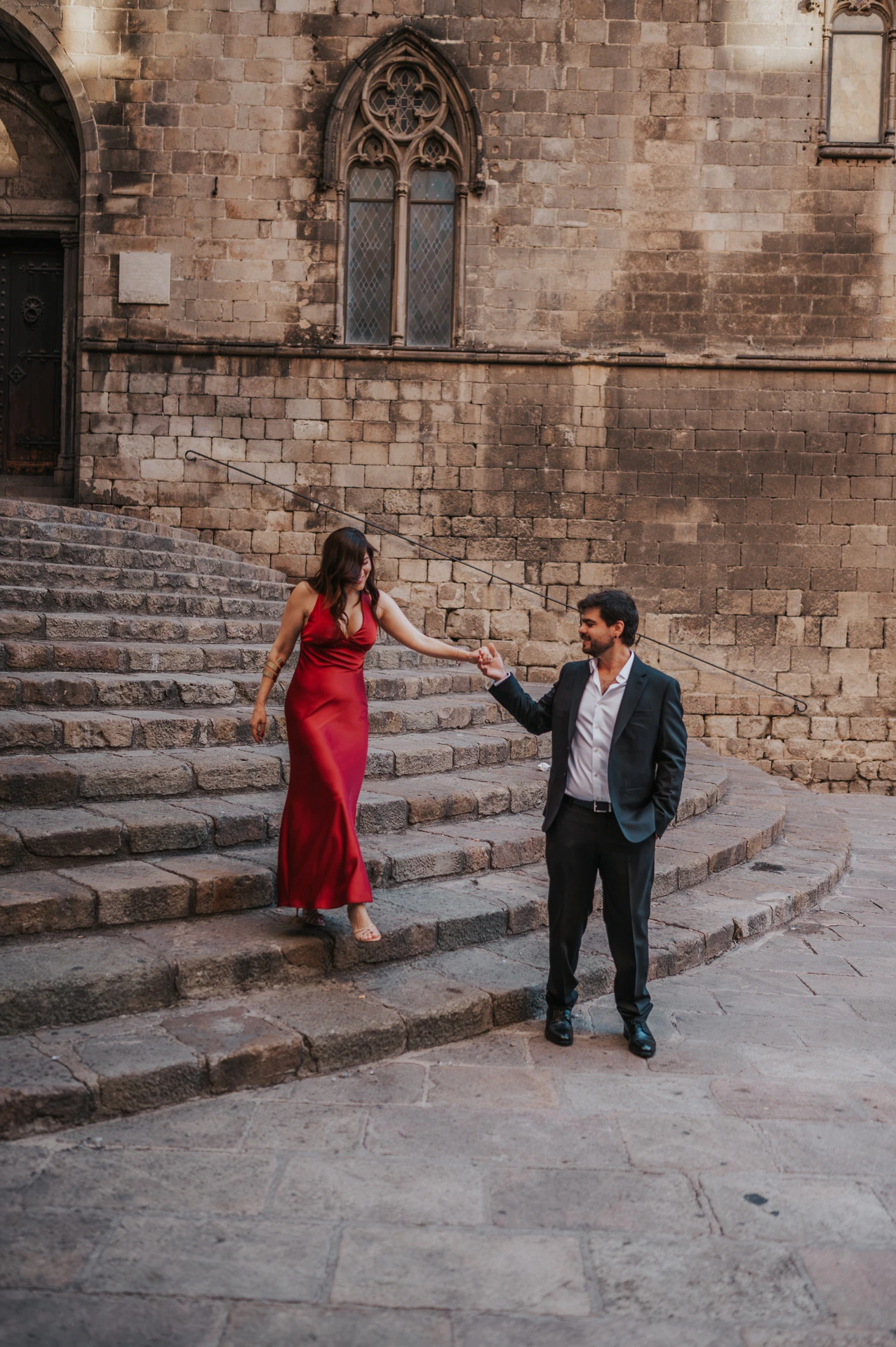 The couple holding hands on the broad stone steps at Plaça del Rei, with historic doors and pointed windows behind them during their engagement photoshoot in Barcelona.