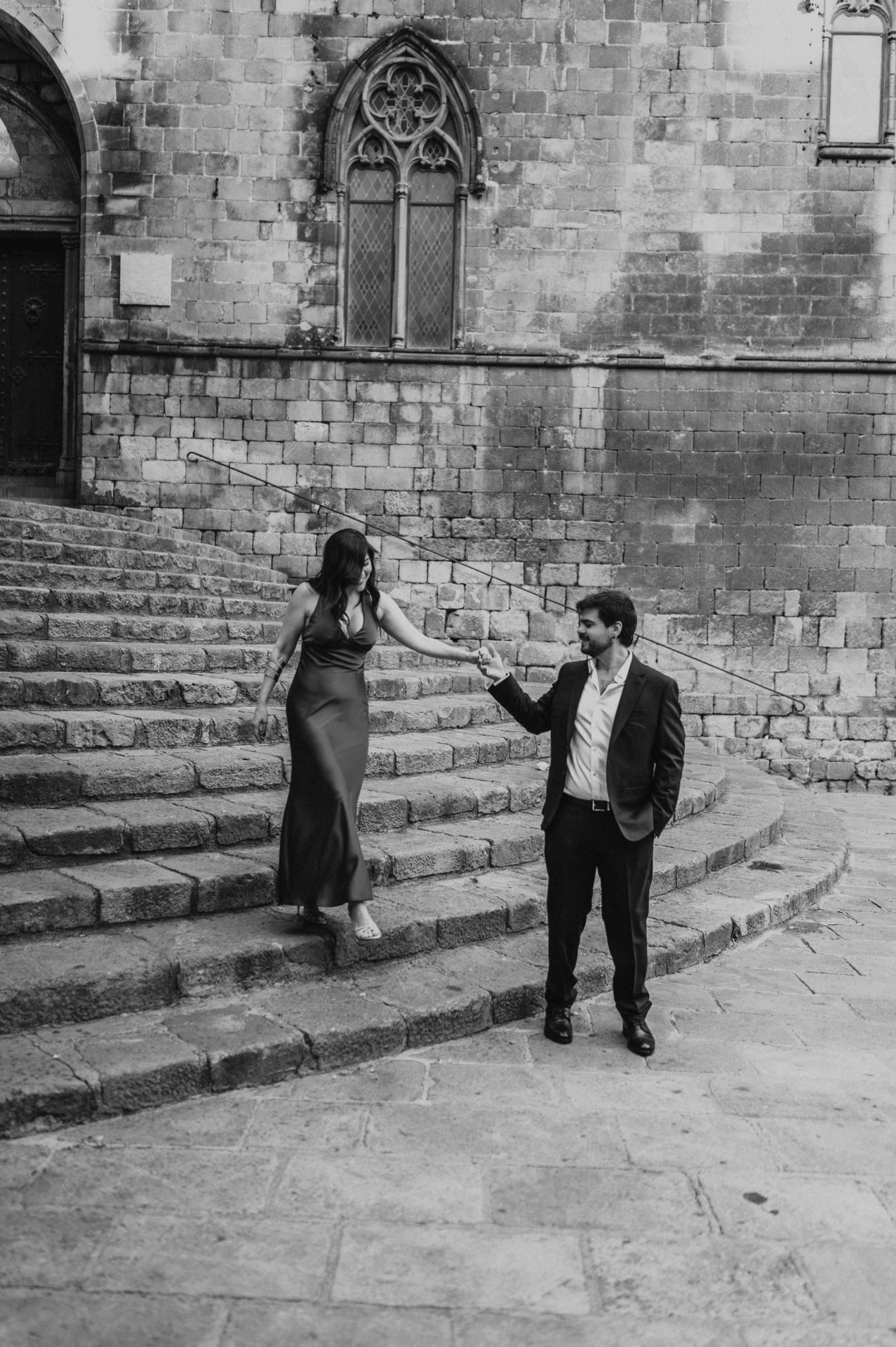 Black-and-white photograph: the couple holding hands on the broad stone steps at Plaça del Rei, with historic doors and pointed windows behind them during their engagement photoshoot in Barcelona.