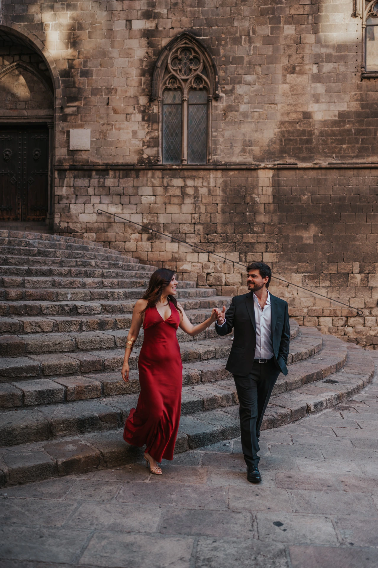The couple holding hands on the broad stone steps at Plaça del Rei, with historic doors and pointed windows behind them during their engagement photoshoot in Barcelona.