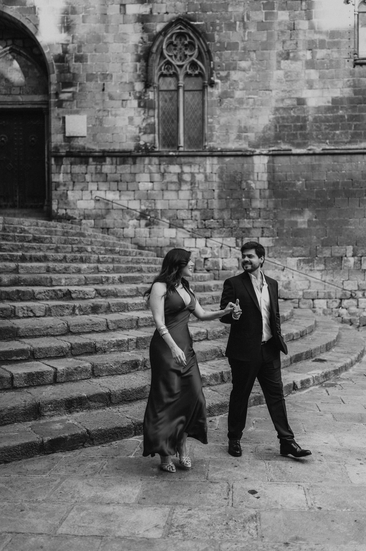 Black-and-white photograph: the couple holding hands on the broad stone steps at Plaça del Rei, with historic doors and pointed windows behind them during their engagement photoshoot in Barcelona.