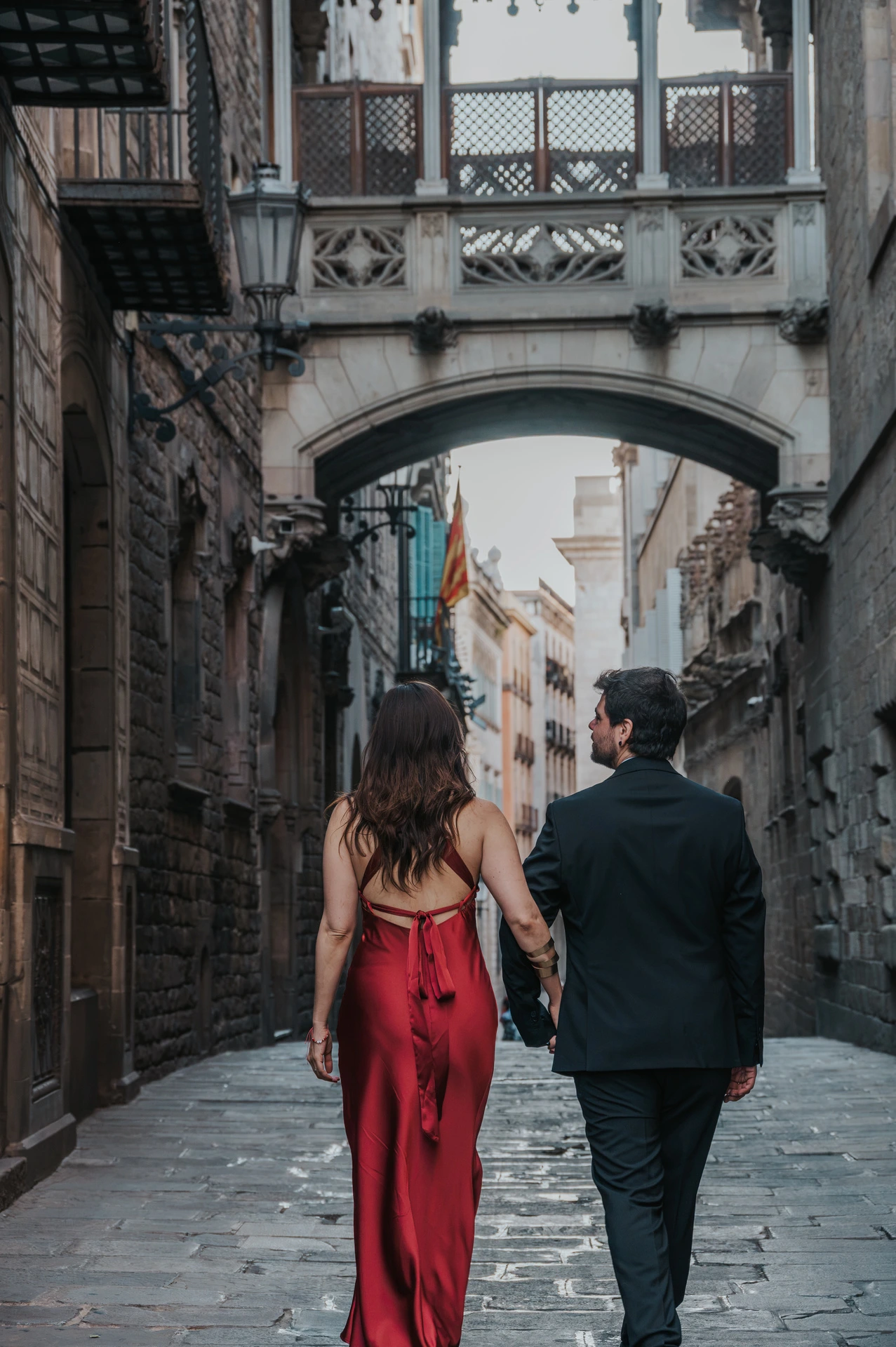 The couple holding hands as they walk away beneath Pont del Bisbe, with the woman’s red dress and the man’s dark suit seen from behind during their engagement photoshoot in Barcelona.