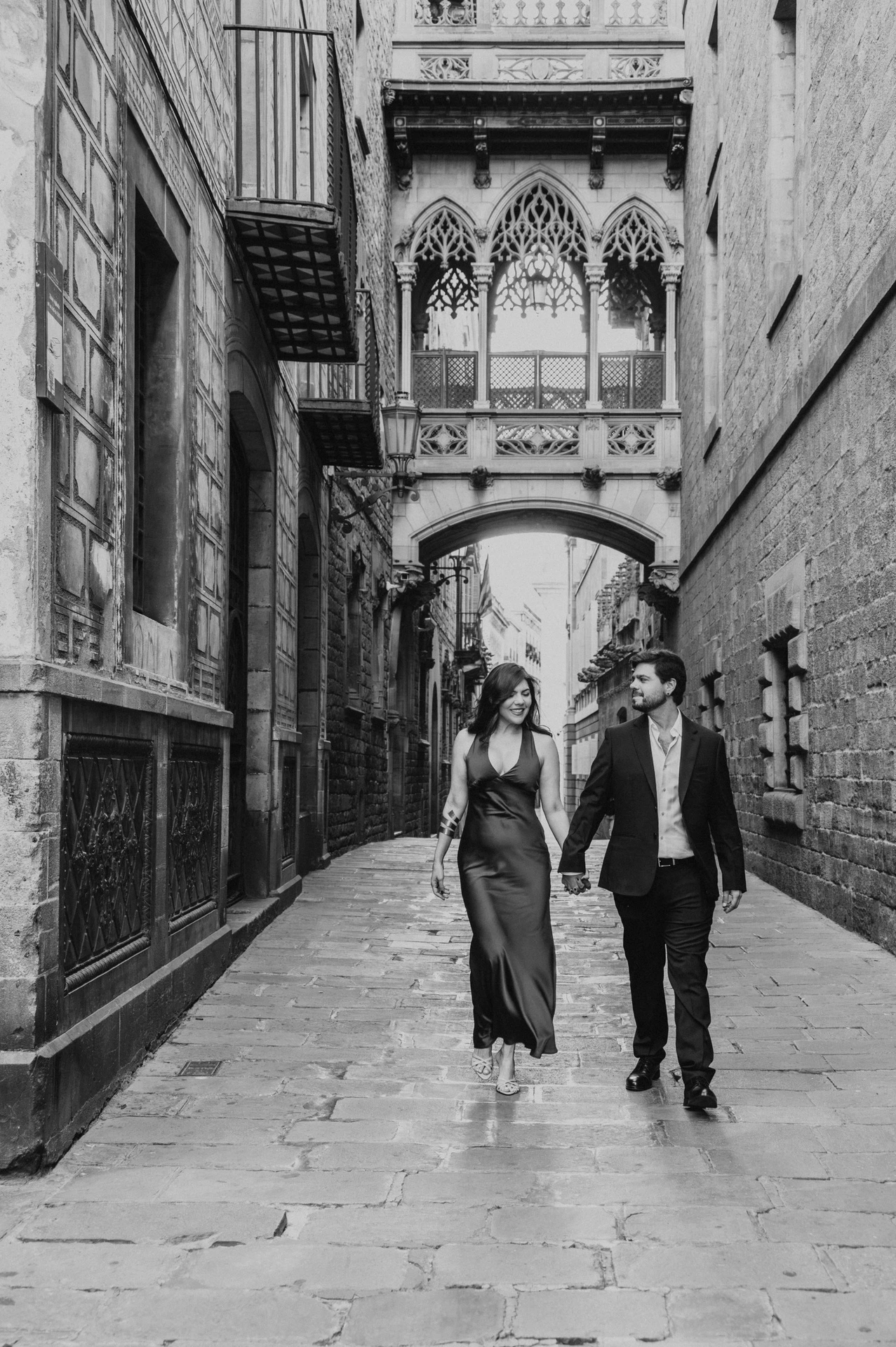 Black-and-white photograph: the couple walking together along the stone lane beneath Pont del Bisbe in the Gothic Quarter during their engagement photoshoot in Barcelona.