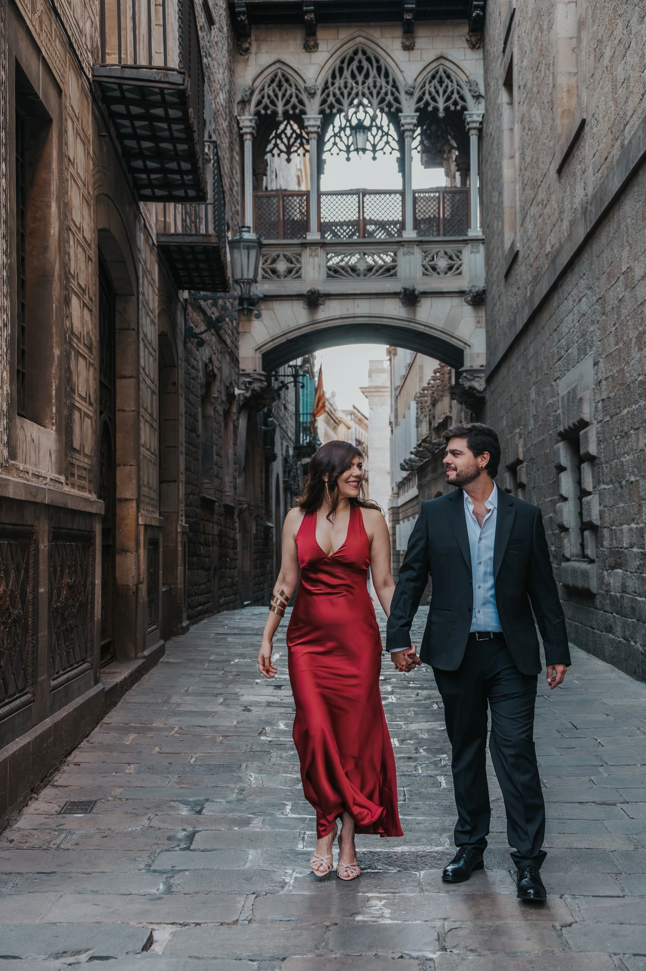 The couple walking together along the stone lane beneath Pont del Bisbe in the Gothic Quarter during their engagement photoshoot in Barcelona.