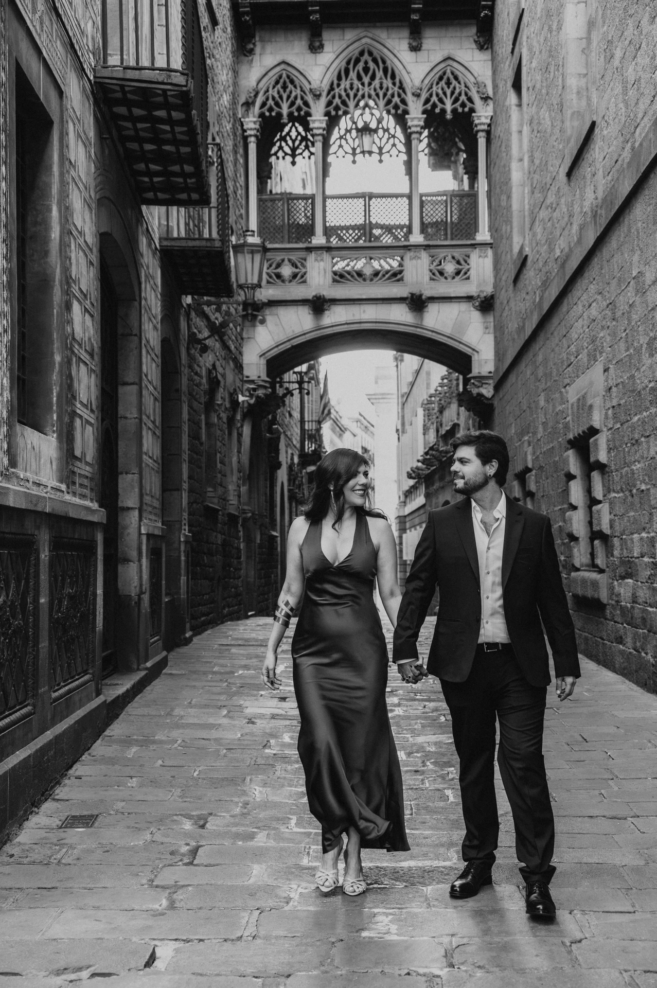Black-and-white photograph: the couple walking together along the stone lane beneath Pont del Bisbe in the Gothic Quarter during their engagement photoshoot in Barcelona.