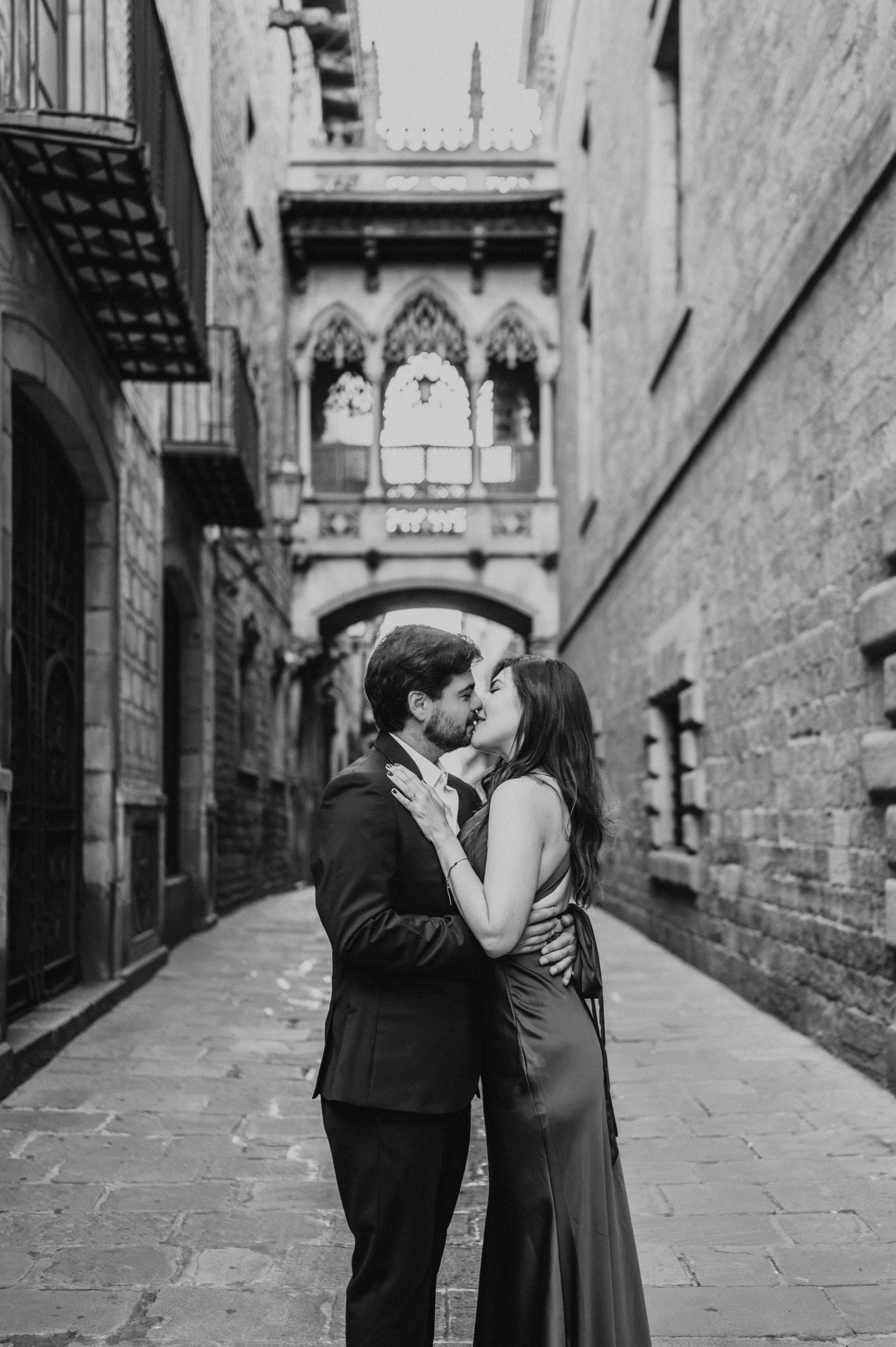 Black-and-white photograph: the couple standing close together beneath Pont del Bisbe, framed by the Gothic Quarter’s old stone buildings during their engagement photoshoot in Barcelona.
