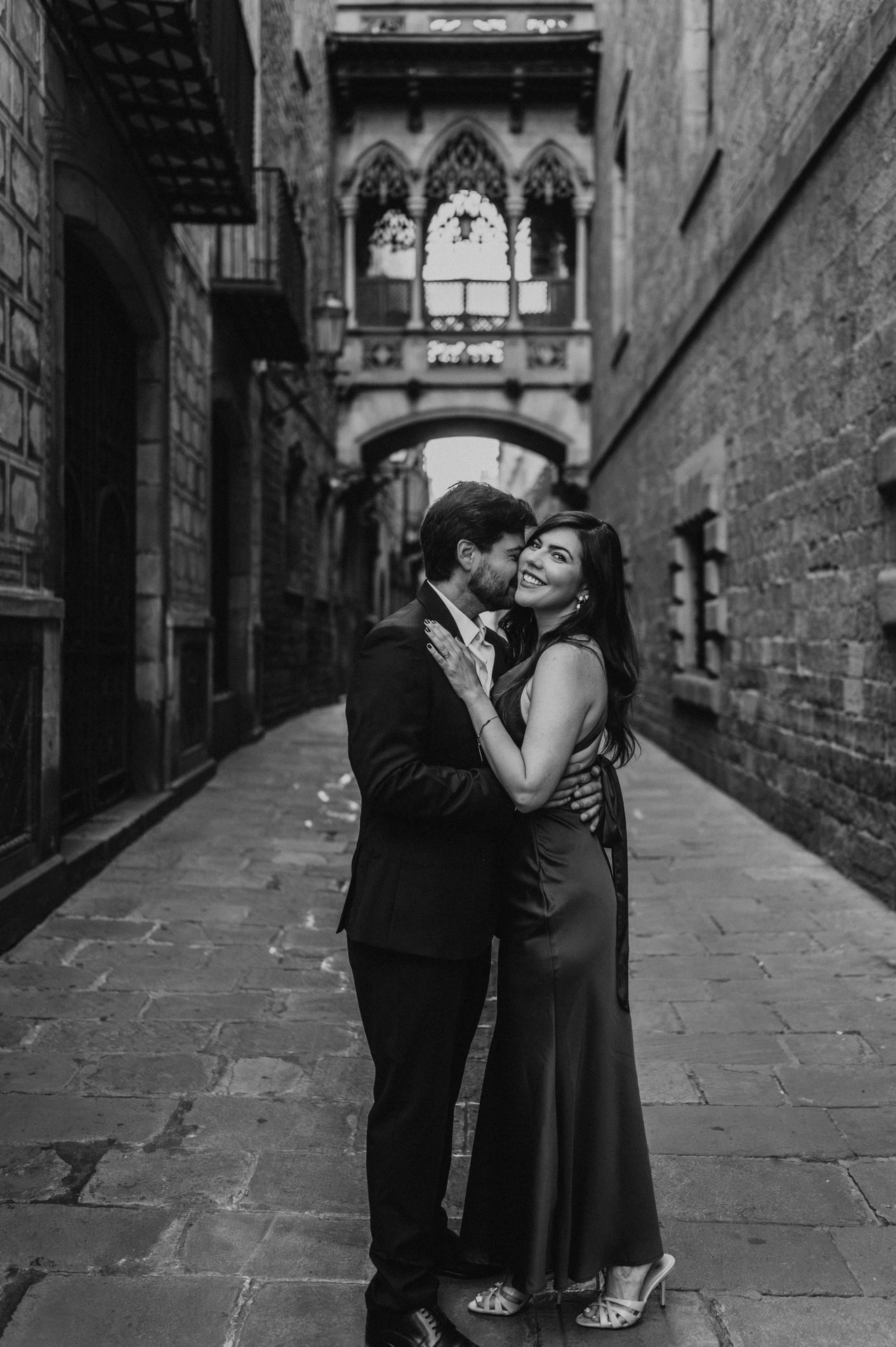 Black-and-white photograph: the couple standing close together beneath Pont del Bisbe, framed by the Gothic Quarter’s old stone buildings during their engagement photoshoot in Barcelona.