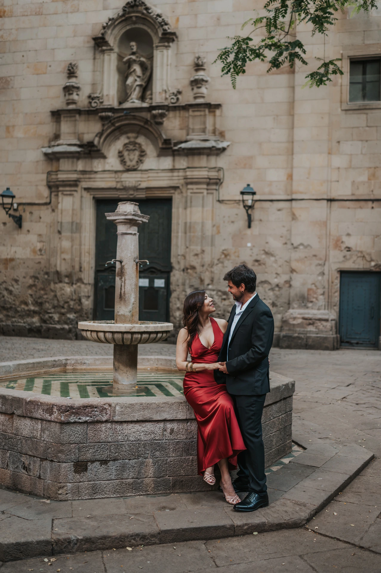 The couple beside the stone fountain in Plaça de Sant Felip Neri, with the church façade behind them during their engagement photoshoot in Barcelona.