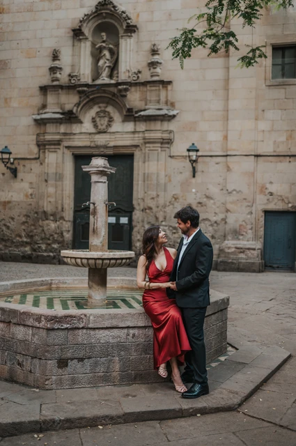 The couple beside the stone fountain in Plaça de Sant Felip Neri, with the church façade behind them during their engagement photoshoot in Barcelona.