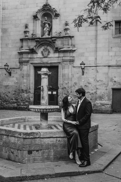 A portrait beside the fountain in Plaça de Sant Felip Neri in black and white | Barcelona engagement photoshoot Black-and-white photograph: the couple beside the stone fountain in Plaça de Sant Felip Neri, with the church façade behind them during their engagement photoshoot in Barcelona.