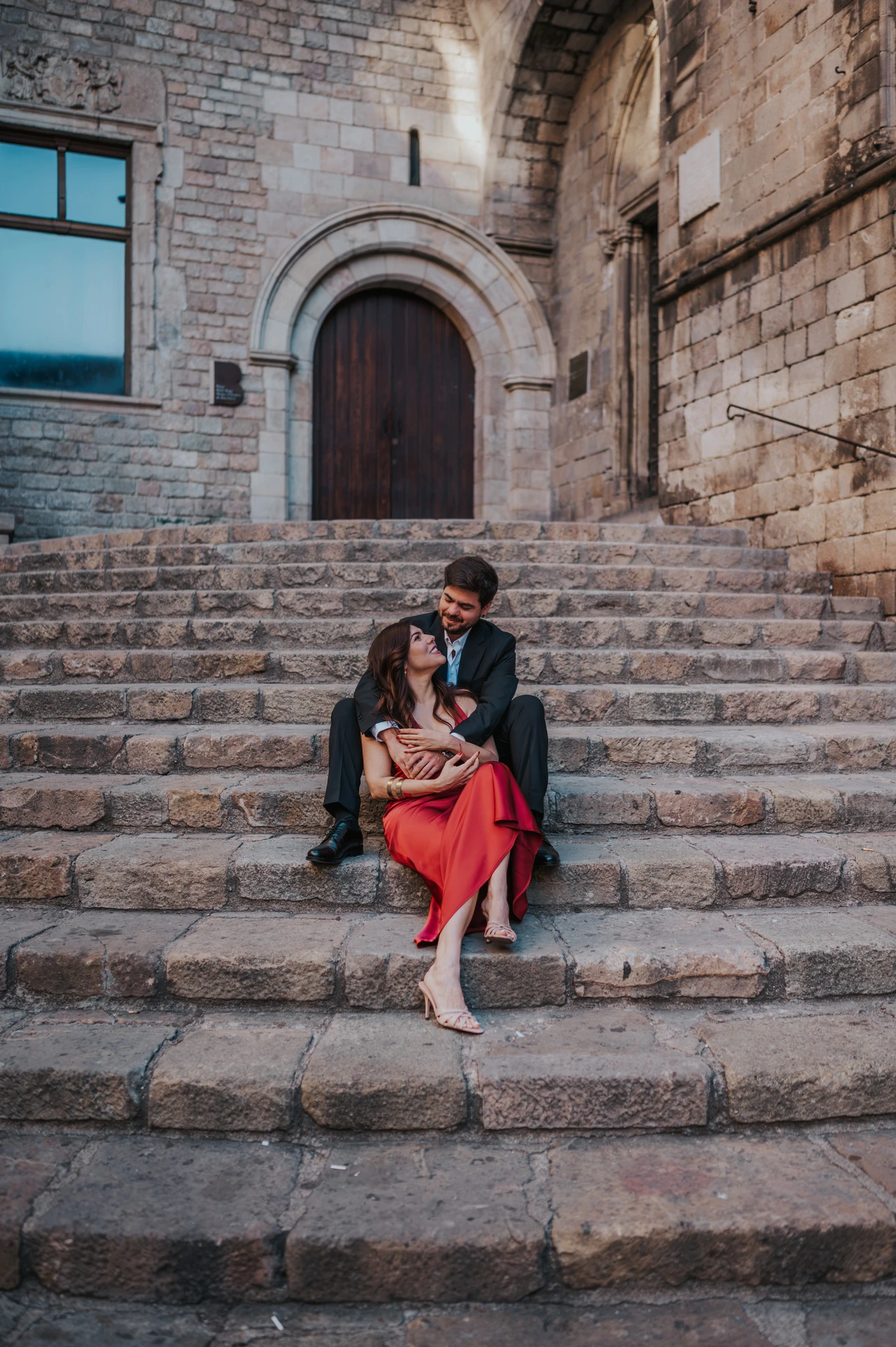The couple sitting close together on the stone steps at Plaça del Rei, beneath an arched doorway during their engagement photoshoot in Barcelona.