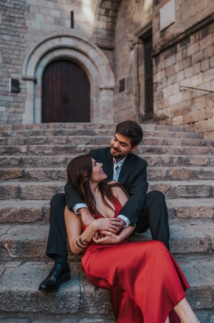 A quiet portrait on the steps of Plaça del Rei | Barcelona engagement photoshoot The couple sitting close together on the stone steps at Plaça del Rei, beneath an arched doorway during their engagement photoshoot in Barcelona.