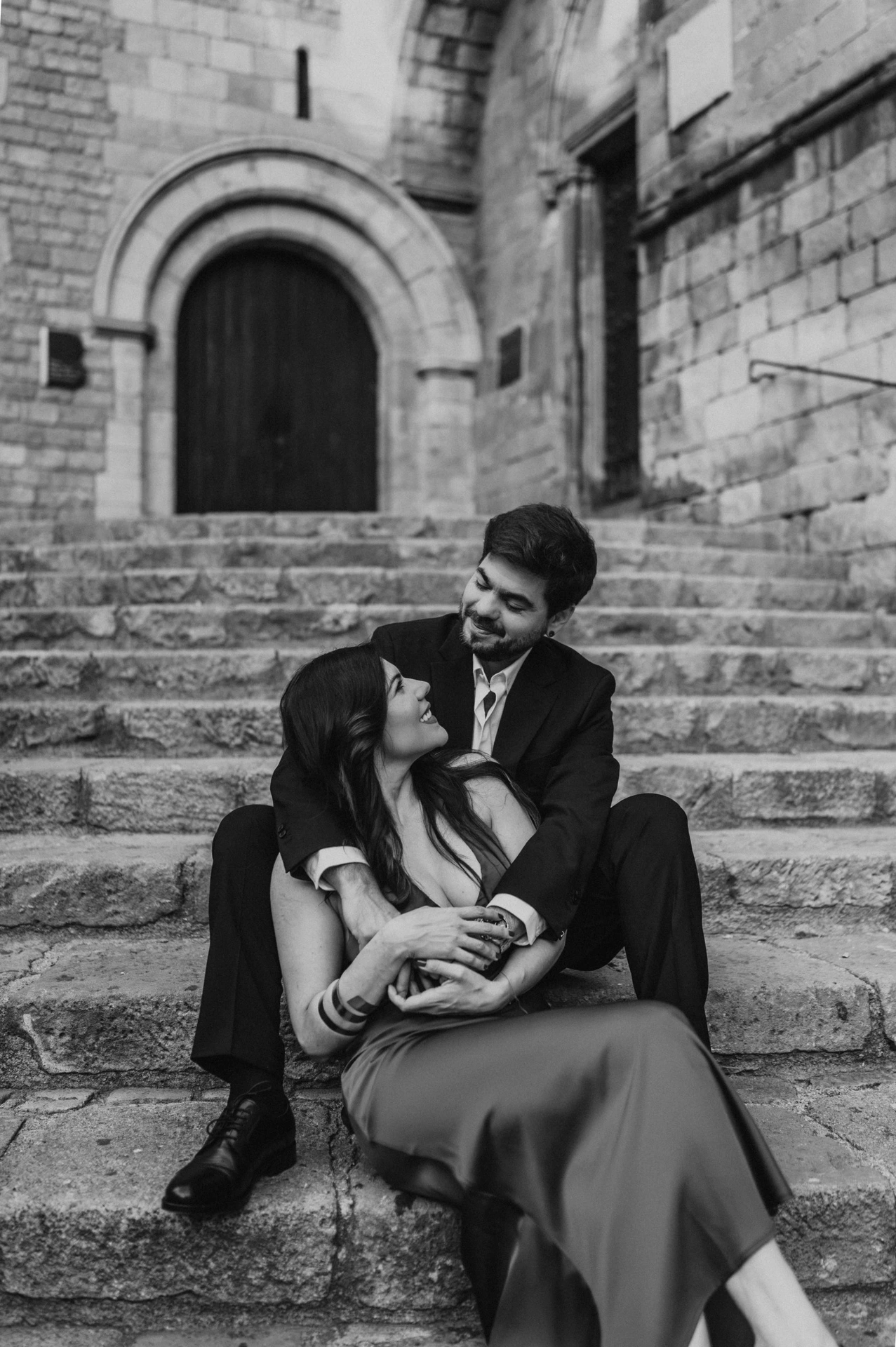 Black-and-white photograph: the couple sitting close together on the stone steps at Plaça del Rei, beneath an arched doorway during their engagement photoshoot in Barcelona.