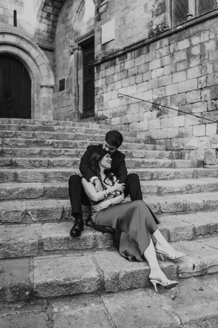 A quiet portrait on the steps of Plaça del Rei in black and white | Barcelona engagement photoshoot Black-and-white photograph: the couple sitting close together on the stone steps at Plaça del Rei, beneath an arched doorway during their engagement photoshoot in Barcelona.