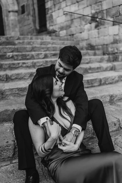 A quiet portrait on the steps of Plaça del Rei in black and white | Barcelona engagement photoshoot Black-and-white photograph: the couple sitting close together on the stone steps at Plaça del Rei, beneath an arched doorway during their engagement photoshoot in Barcelona.